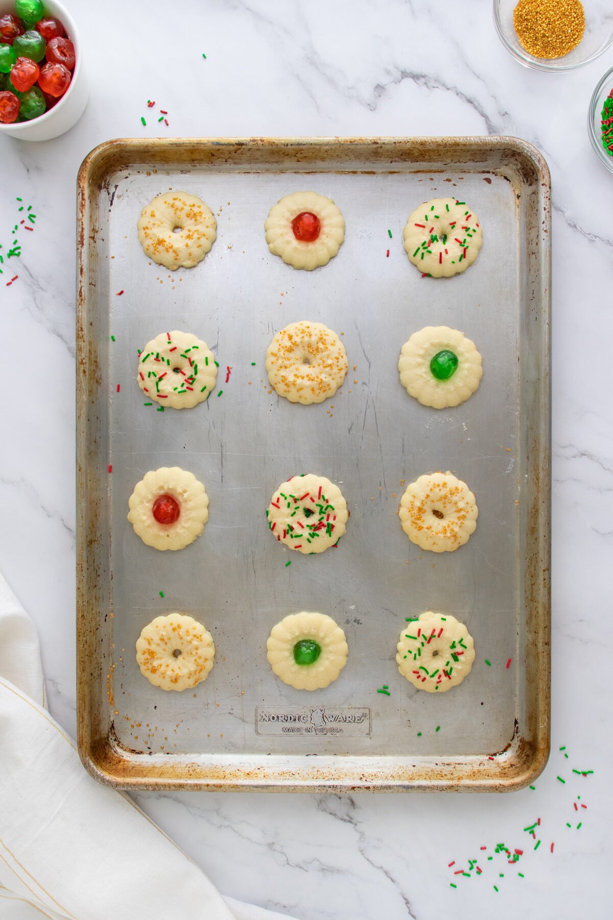 A baking sheet with twelve unbaked cookies, some topped with red or green cherries, others sprinkled with colored sprinkles or crushed nuts, on a marble countertop with jars of sprinkles and cherries nearby.
