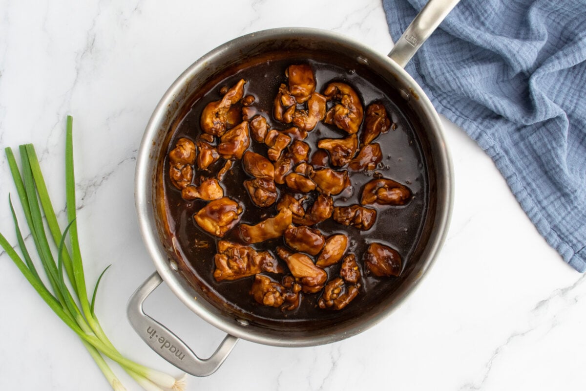 A stainless steel pan filled with pieces of chicken in a dark brown sauce sits on a white marble surface next to three green onions and a folded blue cloth napkin.