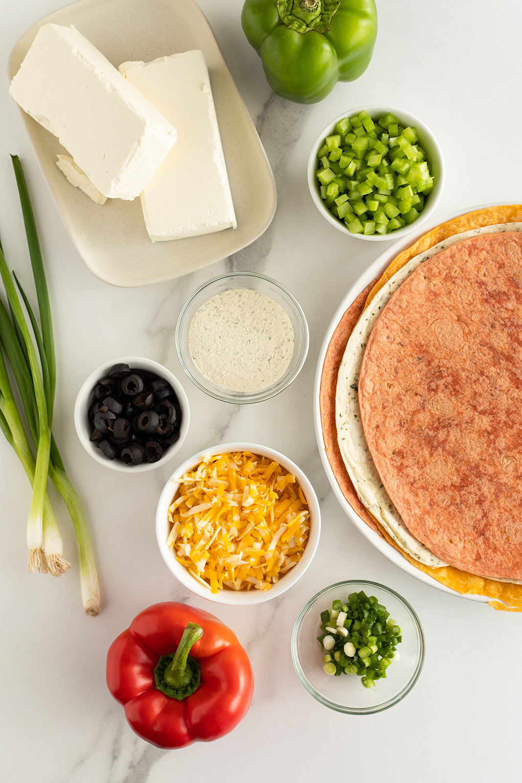 An overhead view of assorted ingredients on a white surface: tortillas, cream cheese, shredded cheese, sliced black olives, chopped green onions, diced celery, a green bell pepper, a red bell pepper, and a bowl of seasoning.