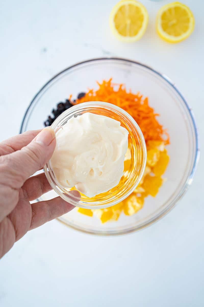 A hand holds a small bowl of mayonnaise over a larger bowl containing shredded carrots, chopped fruit, and raisins; two lemon halves are in the background.