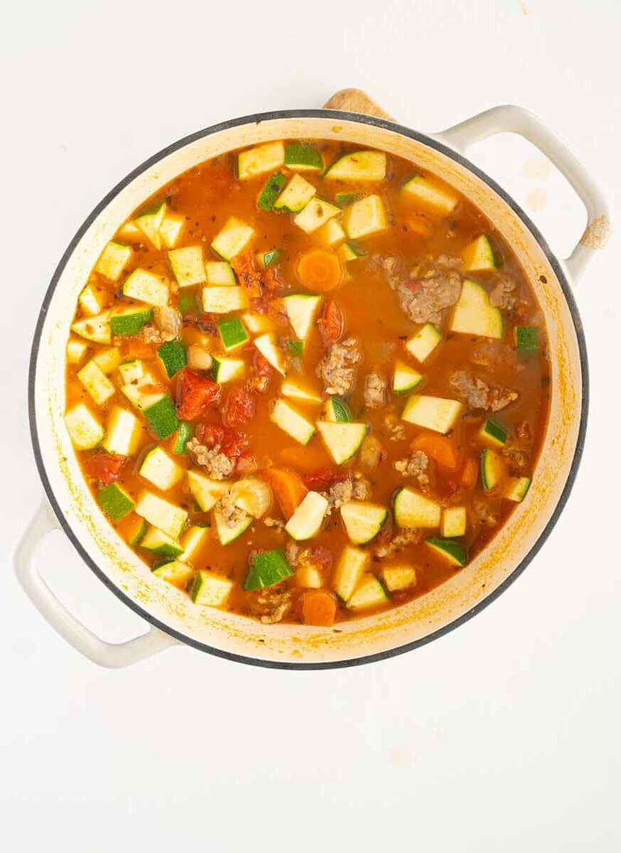 A white pot filled with chunky soup containing pieces of zucchini, tomato, carrots, ground meat, and broth, viewed from above on a white background.