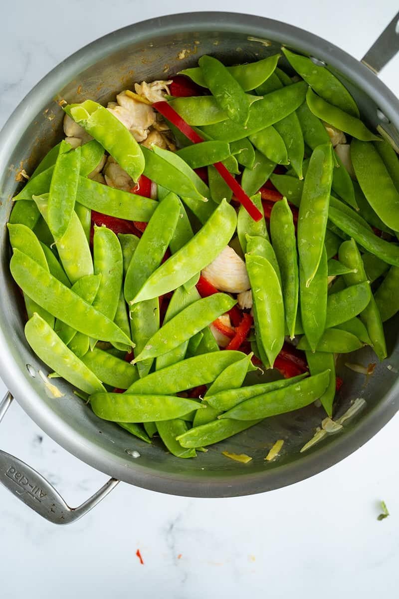 A stainless steel pan filled with fresh snow peas, sliced red bell peppers, and pieces of cooked chicken, all sitting on a white marble surface.