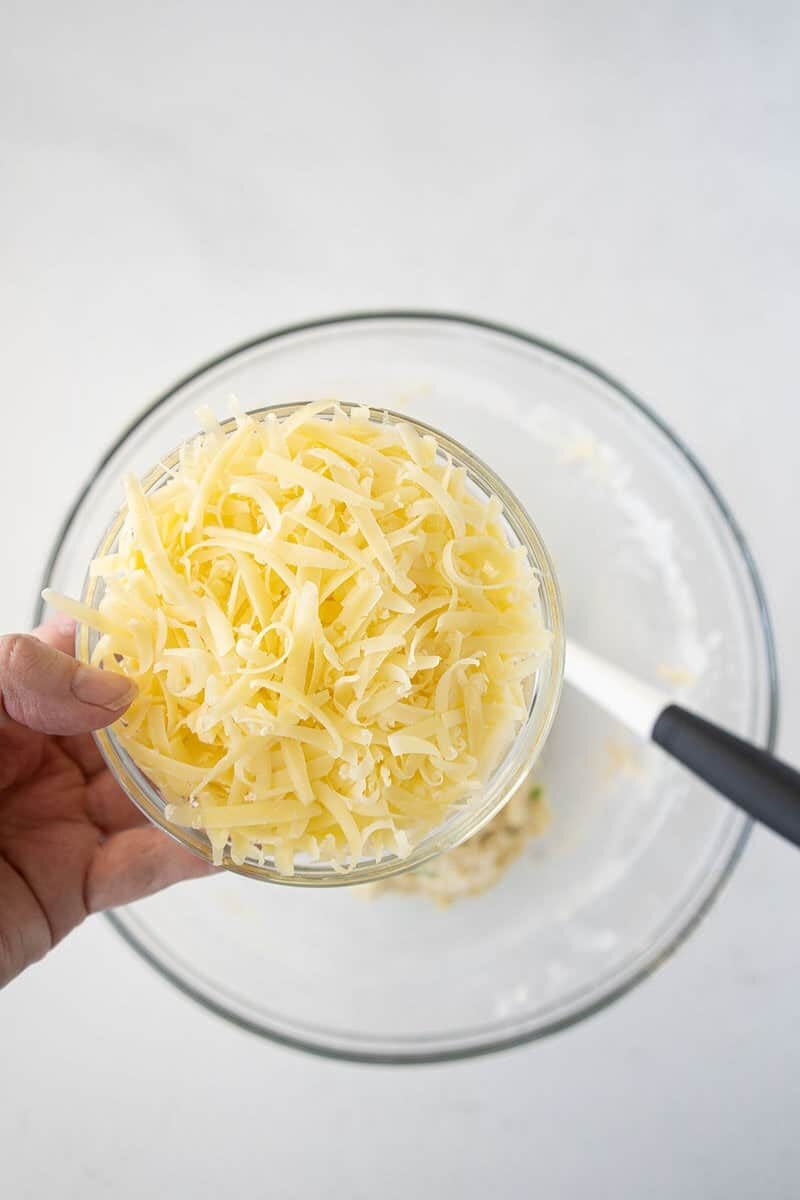 A hand holding a clear glass bowl filled with shredded cheese above a large empty glass mixing bowl with a spatula inside.