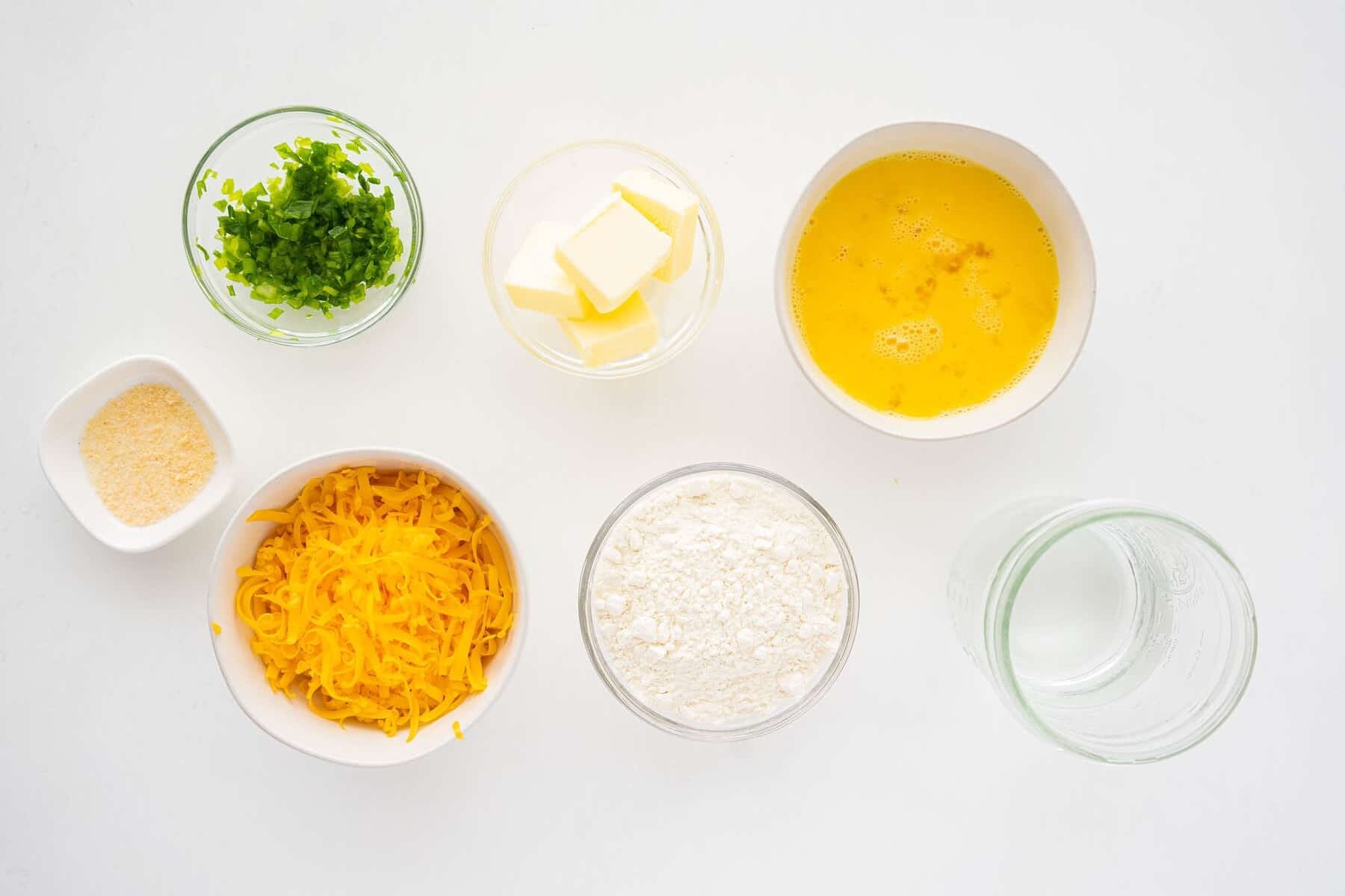 Seven bowls containing ingredients arranged on a white surface: chopped green peppers, butter cubes, beaten eggs, grated cheese, flour, water, and a small bowl of seasoning.