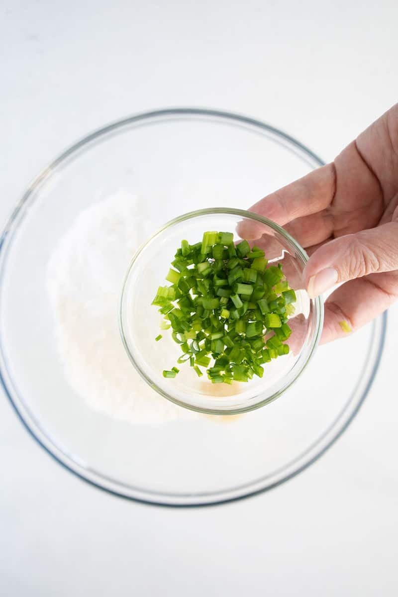 A hand holds a small glass bowl filled with chopped green onions above a larger bowl containing flour on a white surface.
