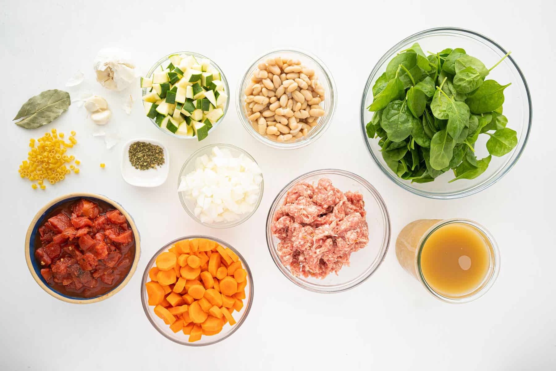 A variety of soup ingredients arranged in bowls on a white surface, including spinach, white beans, diced zucchini, chopped onions, minced garlic, ground meat, diced tomatoes, sliced carrots, broth, dry pasta, and seasonings.