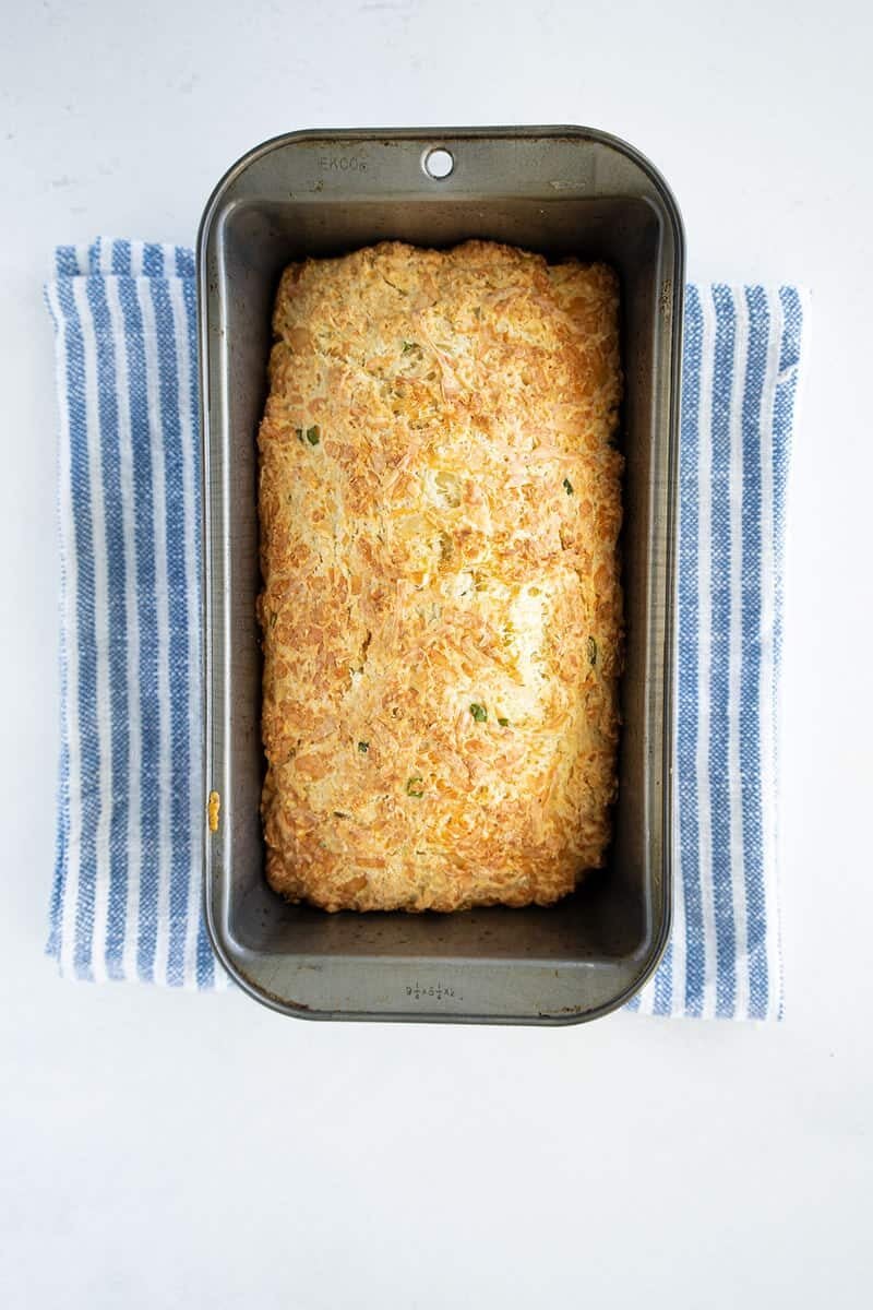 A golden-brown loaf of bread with herbs baked in a metal pan, resting on a blue and white striped cloth against a white background.
