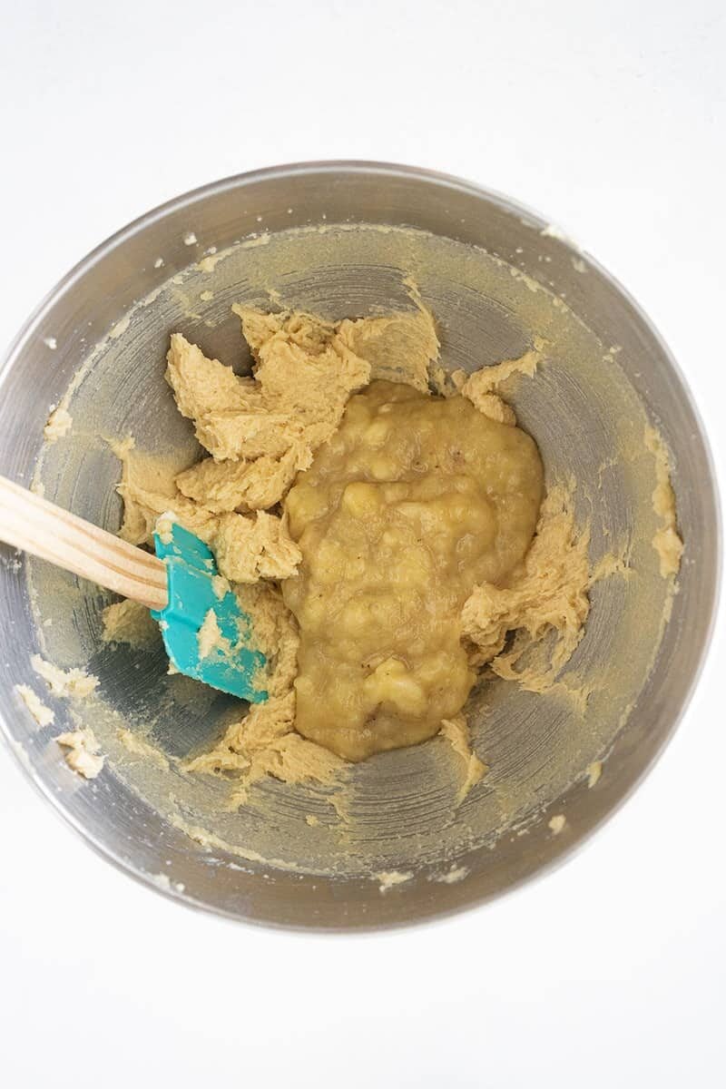 A metal mixing bowl containing creamed dough and mashed bananas, with a teal spatula resting on the side, ready for mixing.