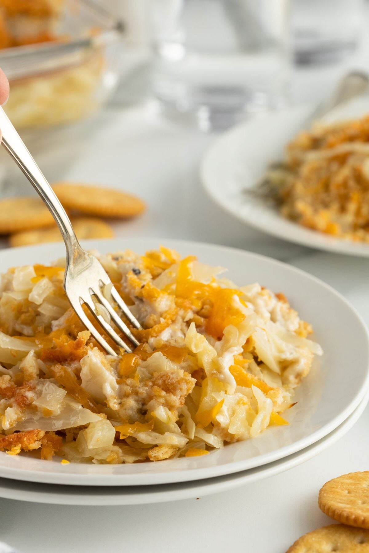 A fork digging into a serving of cheesy casserole on a white plate, with crackers and another plate of casserole in the background on a light-colored table.