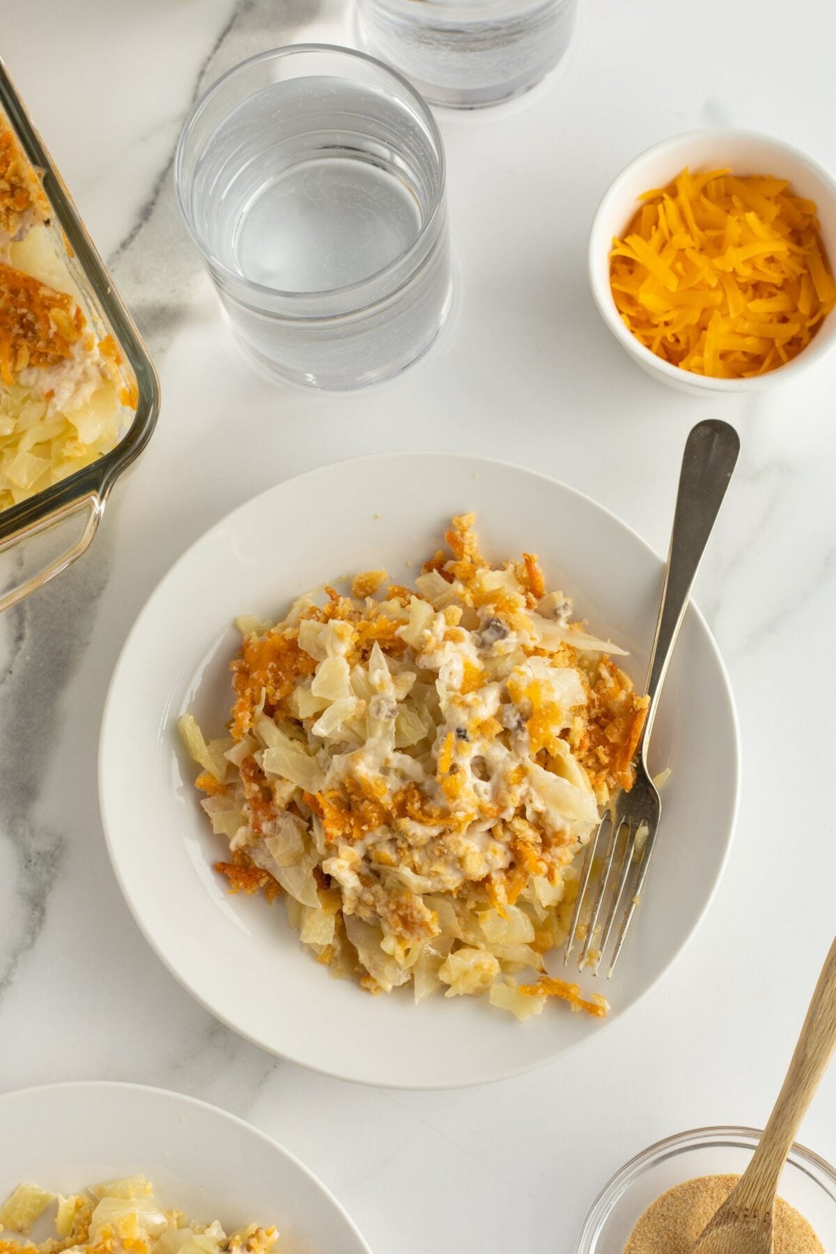 A plate of cheesy potato casserole sits on a white surface with a fork, next to a glass of water, a bowl of shredded cheddar cheese, and a baking dish of casserole.