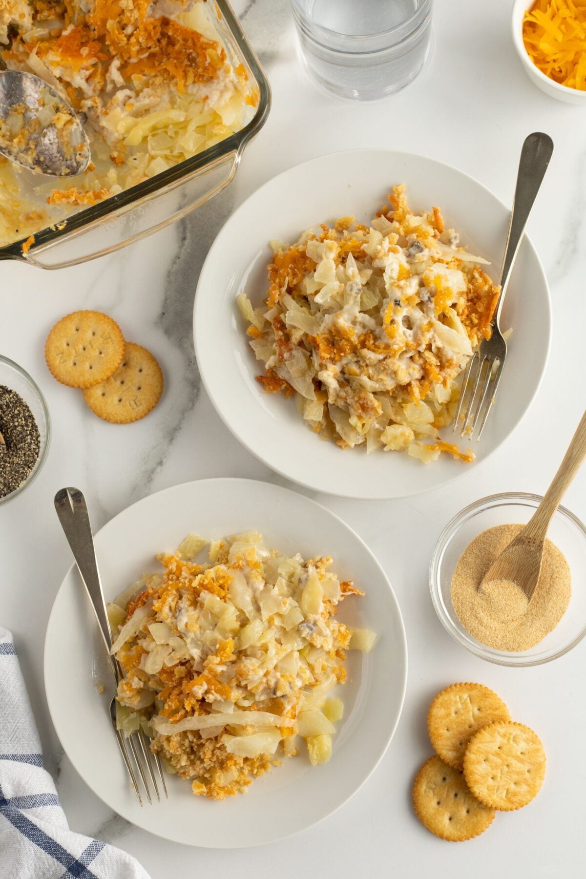 Two white plates each hold a serving of a creamy casserole with crackers on the side. A glass baking dish with more casserole, Ritz crackers, shredded cheese, and seasonings are also visible on a white surface.