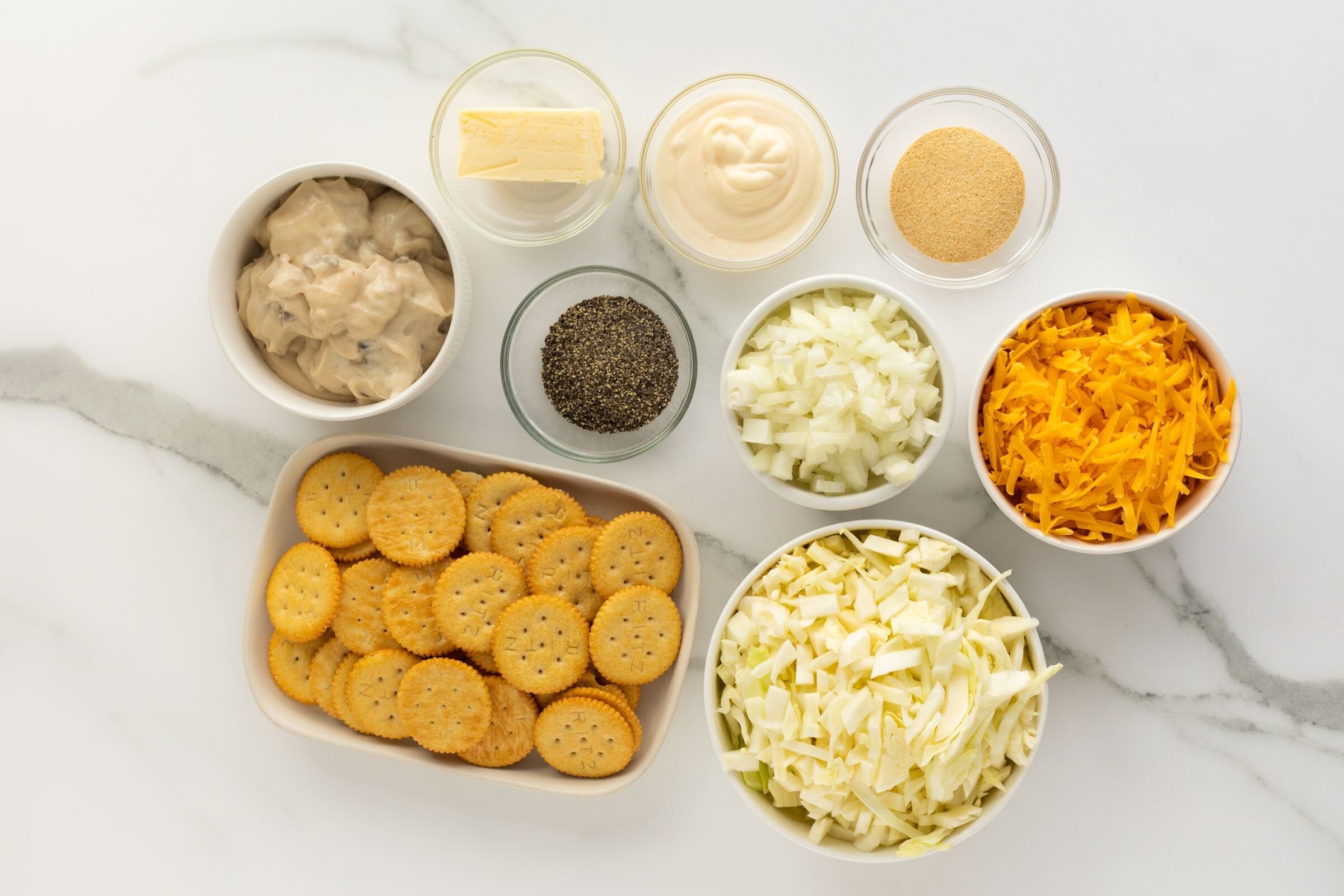 A top-down view of bowls containing round crackers, cream of mushroom soup, butter, black pepper, chopped cabbage, chopped onion, shredded cheddar cheese, mayonnaise, and breadcrumbs on a white surface.