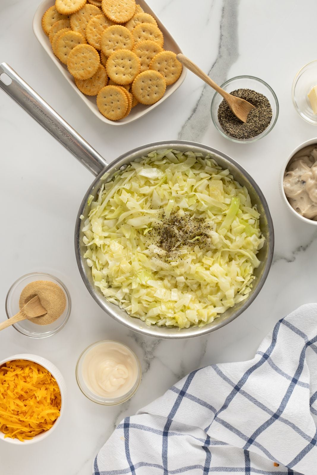 A saucepan filled with chopped cabbage and black pepper sits on a marble countertop, surrounded by shredded cheese, mayonnaise, seasoning, a bowl of crackers, and a blue-and-white checkered kitchen towel.