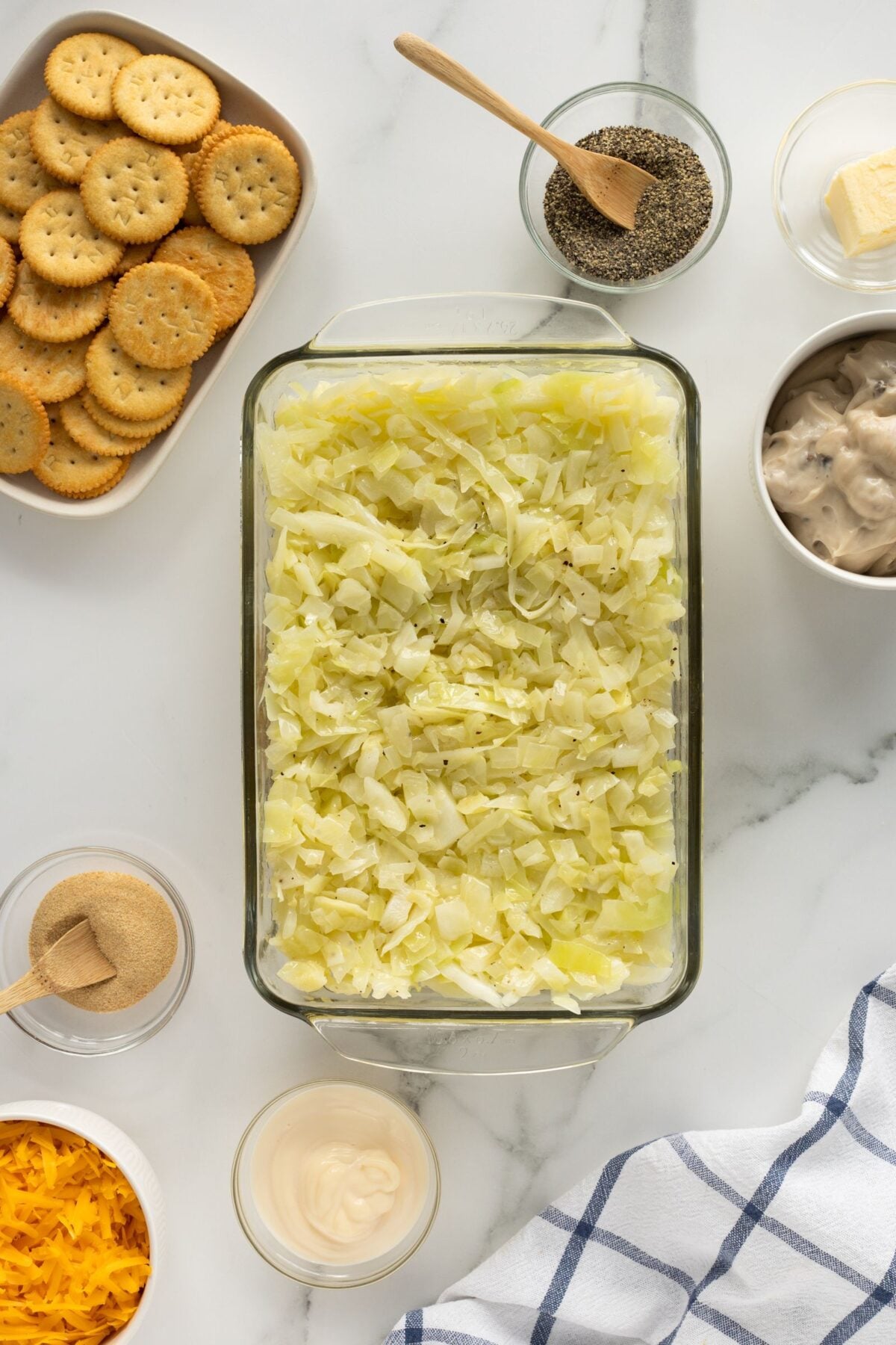 A glass baking dish filled with chopped cabbage is surrounded by small bowls of pepper, butter, shredded cheese, mayonnaise, Ritz crackers, garlic powder, and a can of soup on a marble countertop with a striped towel.