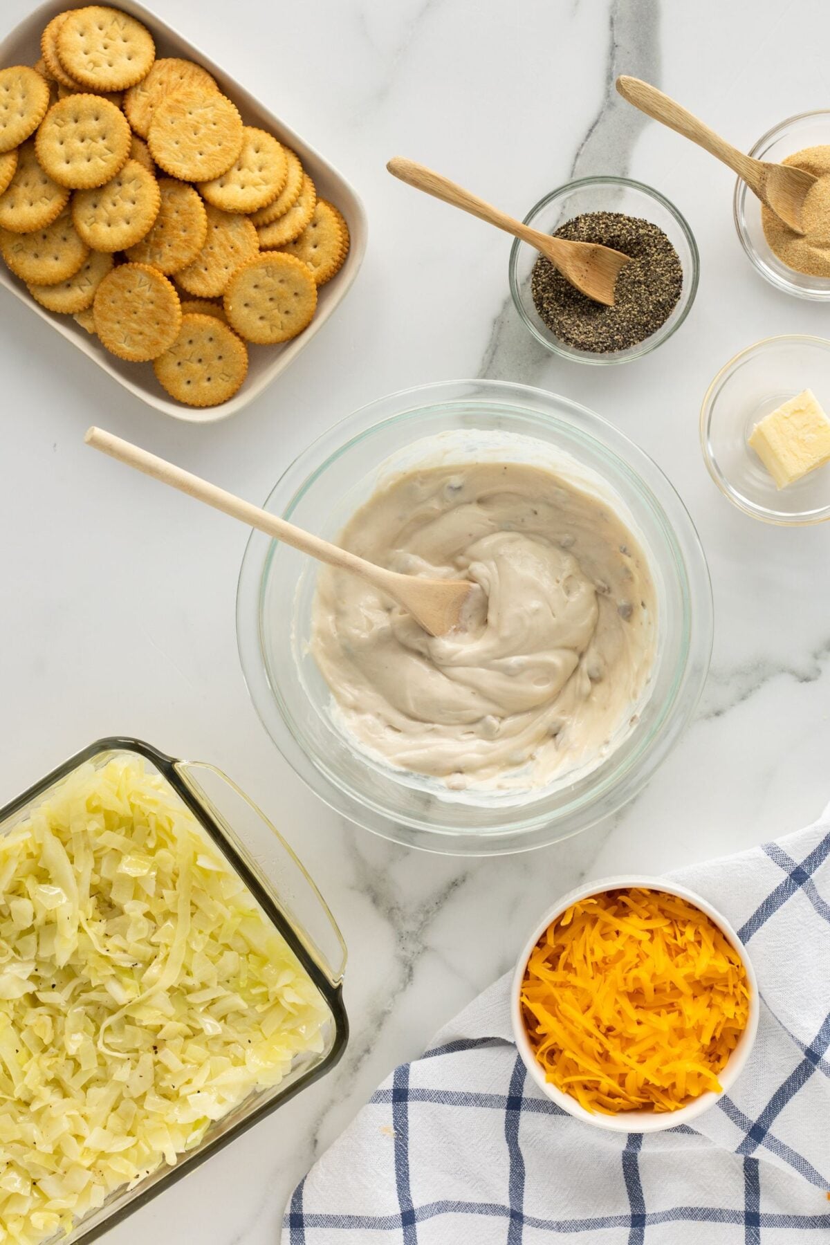 A top-down view of ingredients for a casserole: a bowl of creamy mixture with a wooden spoon, a tray of shredded cabbage, a bowl of shredded cheddar, round crackers, butter, ground pepper, and a blue checkered towel.