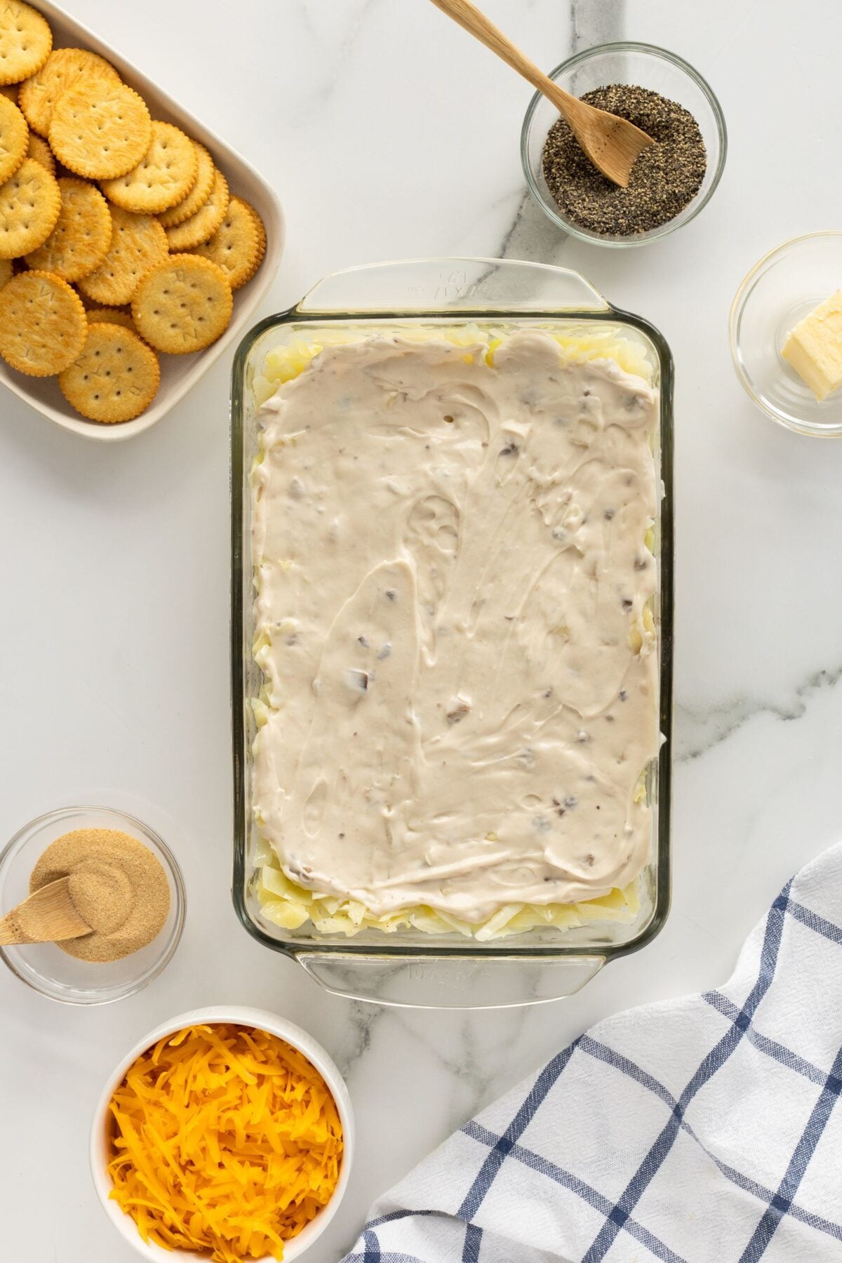 A glass baking dish with a creamy mixture spread on top, surrounded by bowls of shredded cheddar cheese, pepper, a stick of butter, and a plate of round crackers on a white marble surface.