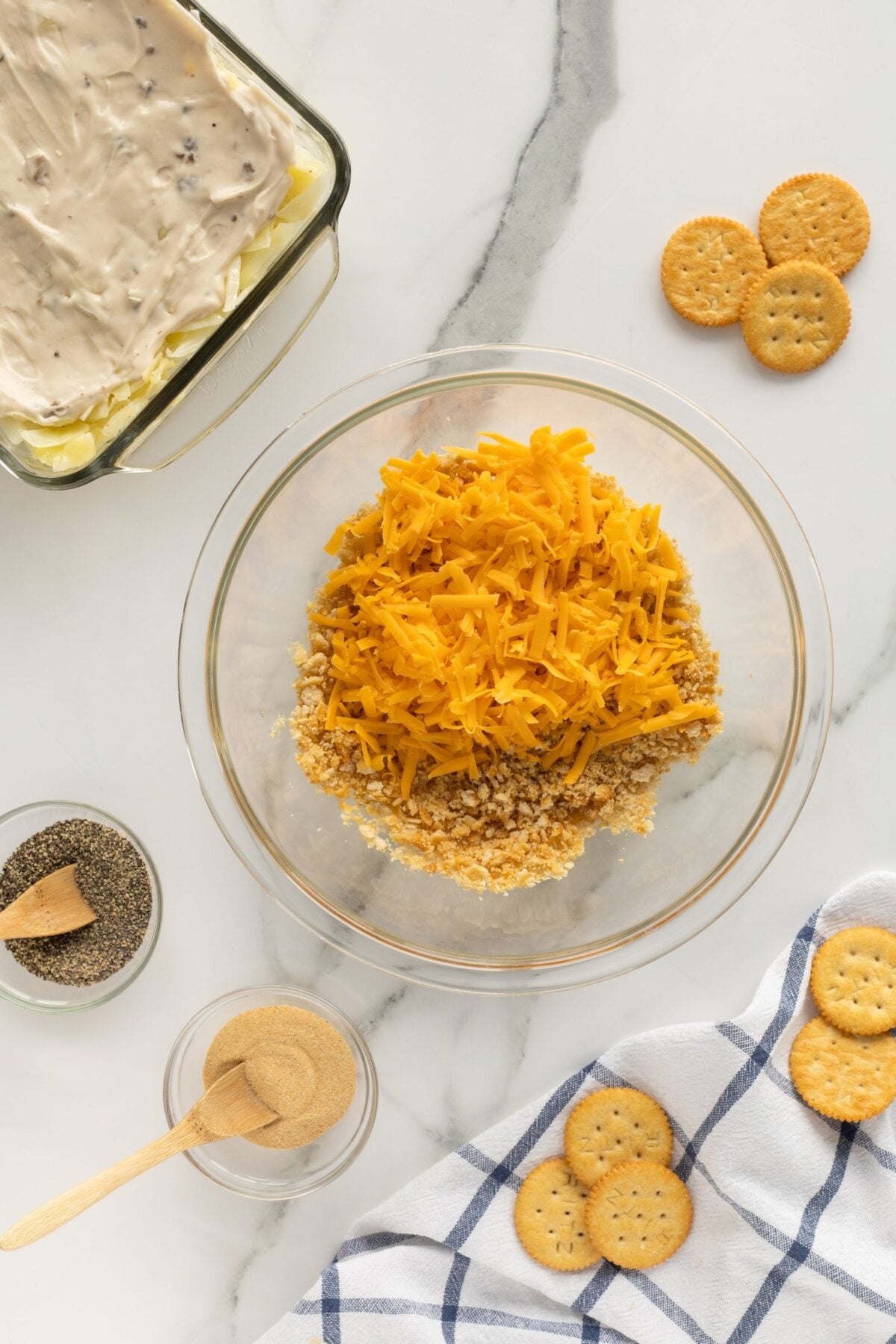 A glass bowl with shredded cheese and crushed crackers sits on a marble counter. Nearby are whole round crackers, a dish with creamy mixture, bowls of pepper and seasoning, and a blue-striped cloth.