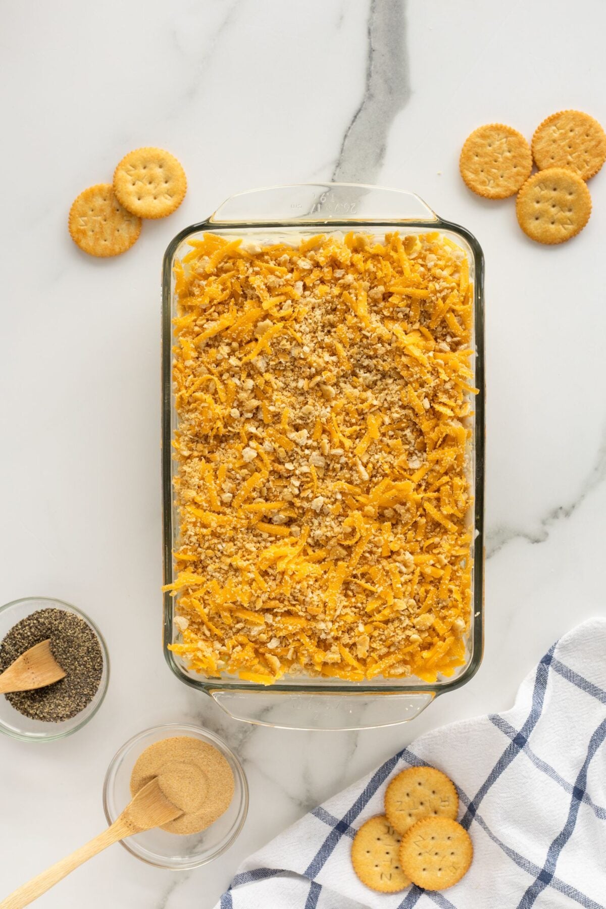 A glass baking dish filled with a casserole topped with shredded cheese and crushed crackers sits on a marble surface, surrounded by round crackers, a bowl of pepper, and a bowl of seasoning next to a striped kitchen towel.