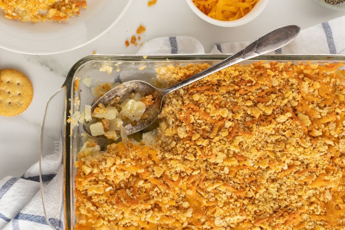 A glass baking dish filled with a golden, crumbly casserole with a spoon scooping out a portion. Ritz crackers, cheese, and a partial serving on a plate are visible nearby on a white surface.