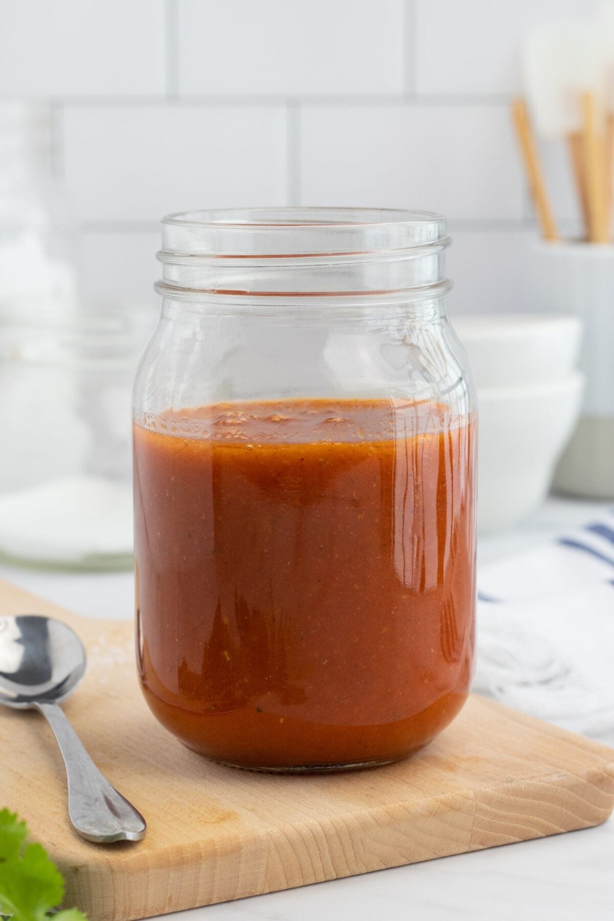 A glass mason jar filled with red taco sauce sits on a wooden cutting board next to a metal spoon; kitchen bowls and utensils are blurred in the background.