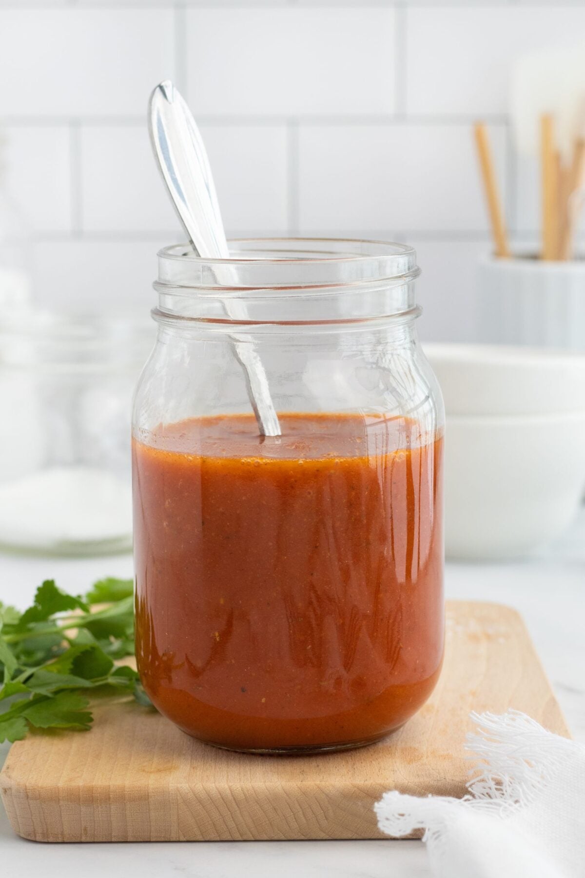 A glass jar filled with red enchilada sauce sits on a wooden board with a metal spoon inside. Fresh cilantro and a white cloth are nearby, and kitchen items are blurred in the background.