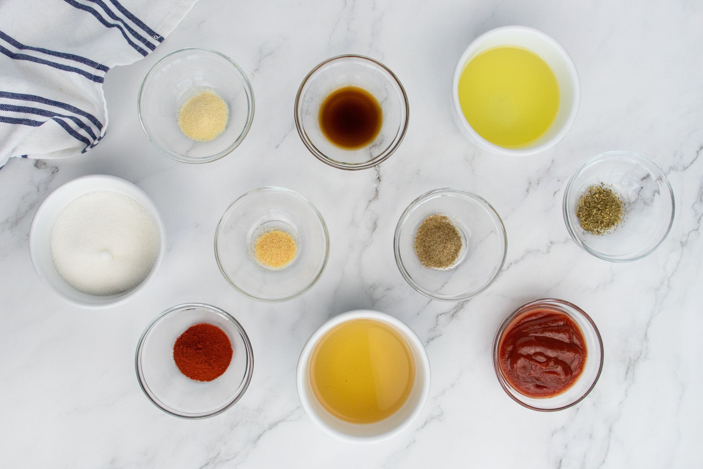 Ten small glass bowls on a marble surface, each containing a different ingredient including olive oil, ketchup, apple cider vinegar, sugar, spices, and seasonings; a striped cloth is at the corner.