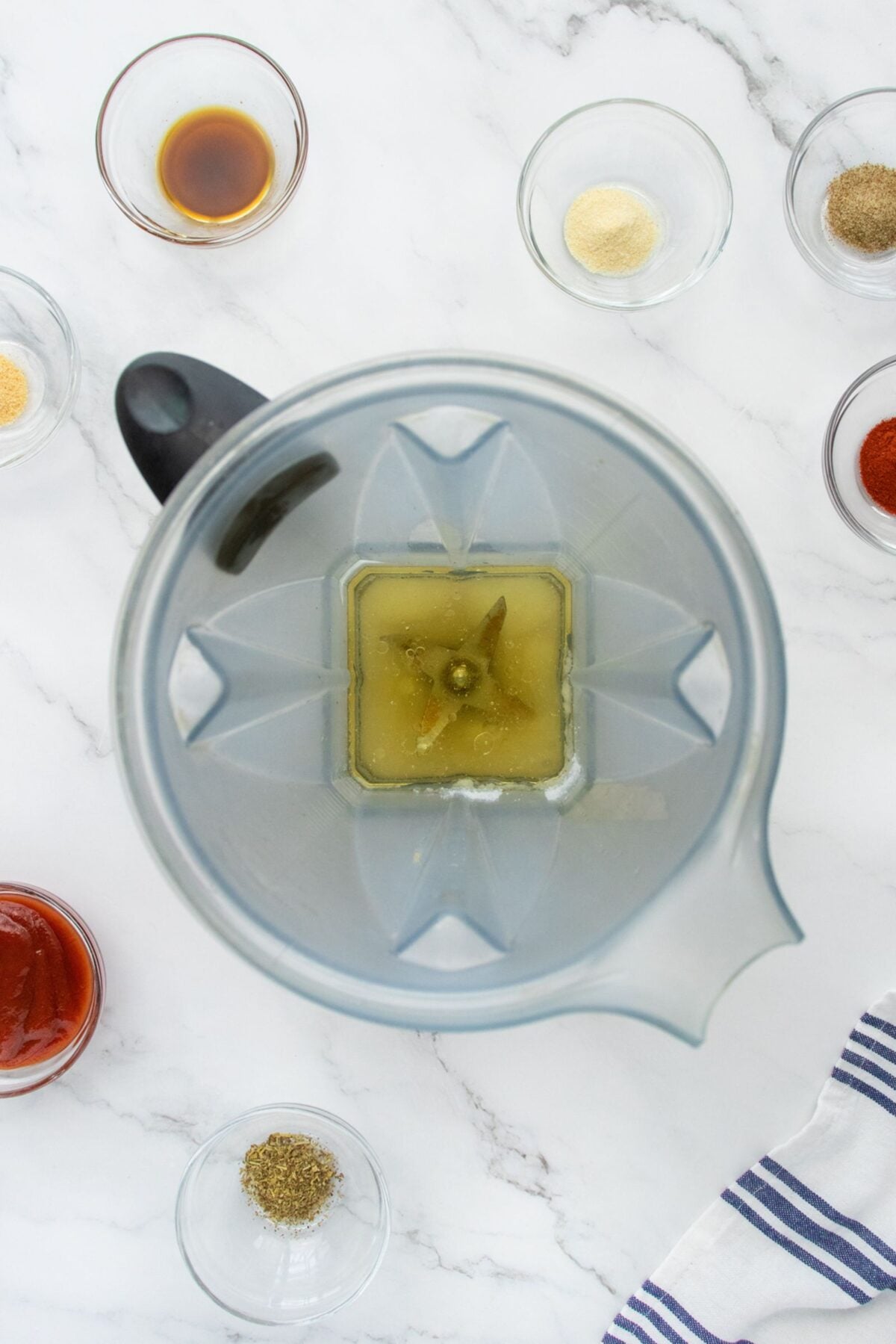 A top-down view of a blender containing greenish liquid and seasonings, surrounded by small bowls holding various spices and sauces on a white marble countertop.