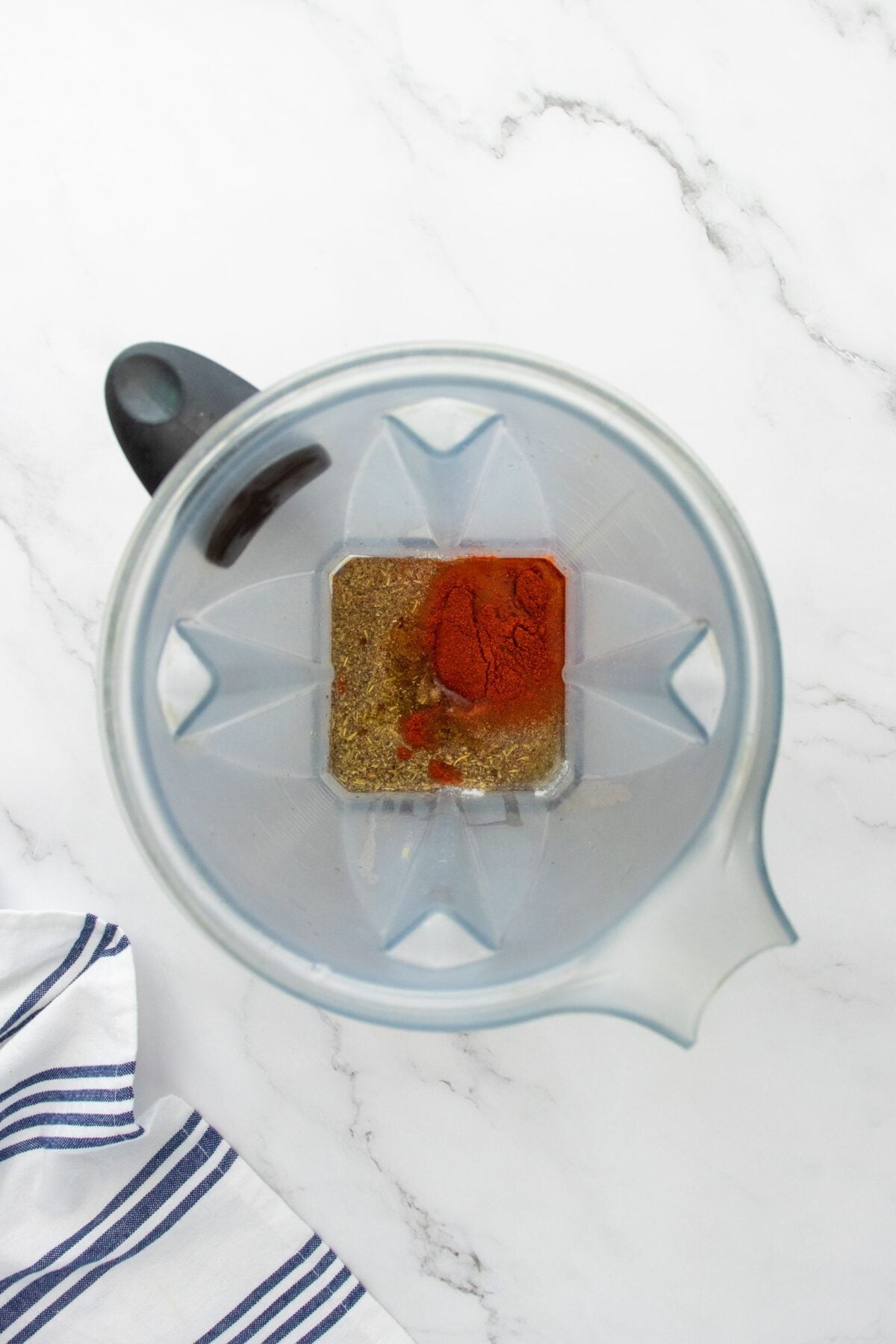 A top-down view of a blender containing various ground spices, including a prominent heap of red spice, on a white marble surface next to a white towel with blue stripes.