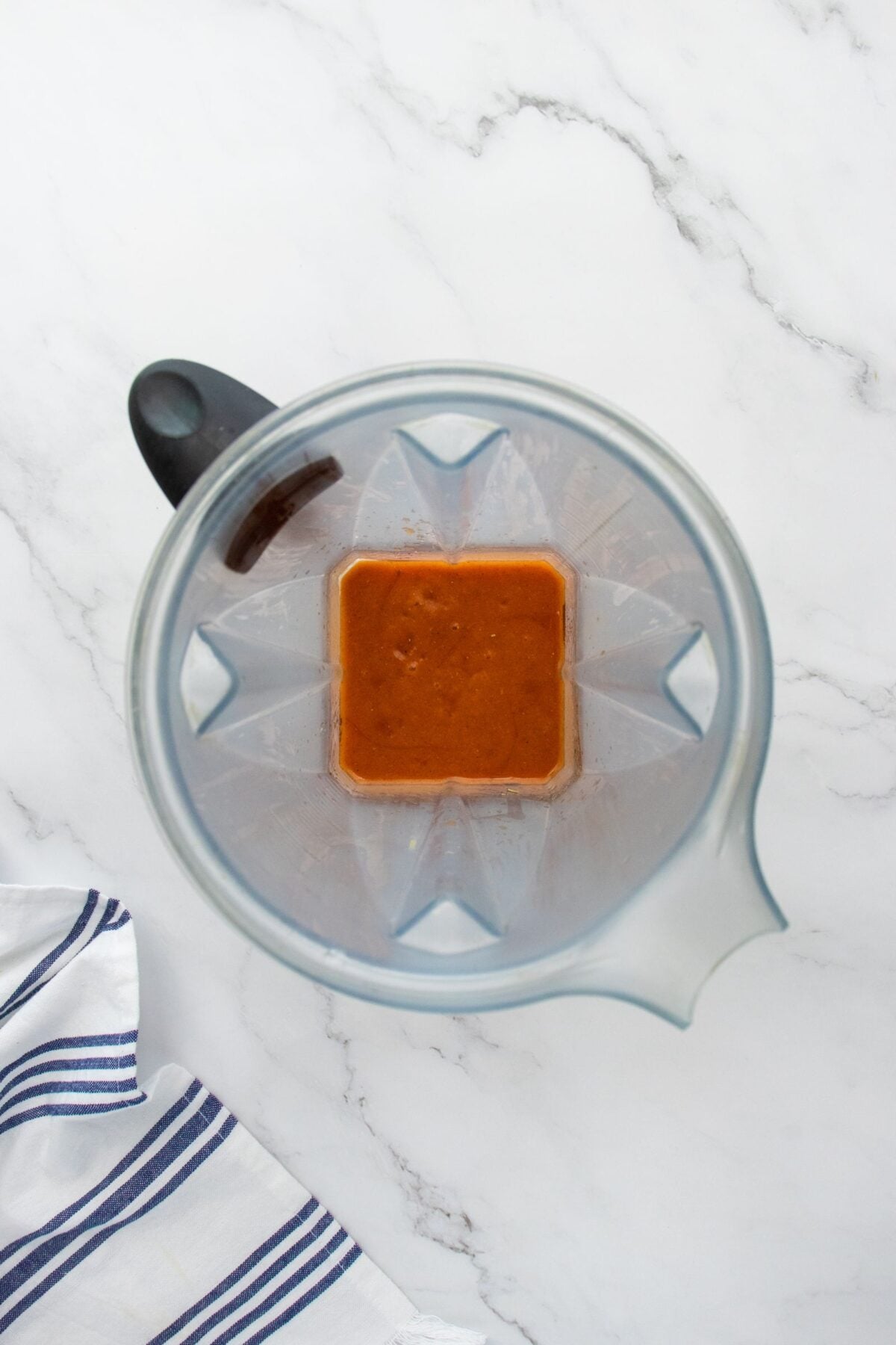 Overhead view of a blender with a reddish-brown sauce inside, placed on a white marble countertop. Part of a white cloth with blue stripes is visible in the lower left corner.