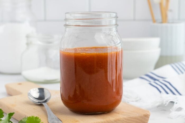 A glass jar filled with red enchilada sauce sits on a wooden cutting board next to a metal spoon, with bowls and kitchen items blurred in the background.