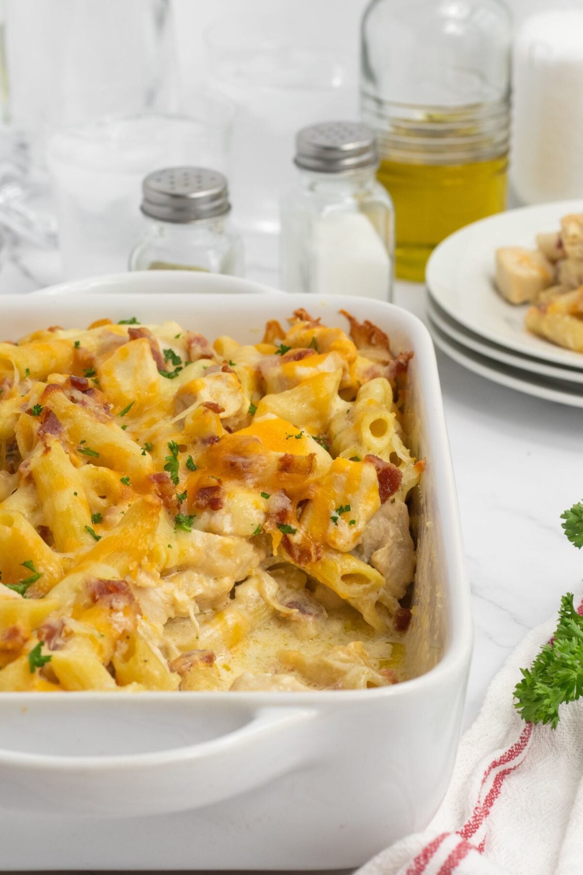 A close-up of a baked pasta casserole topped with melted cheese and herbs in a white dish. In the background are plates, glasses, salt and pepper shakers, and a jar of olive oil on a white counter.