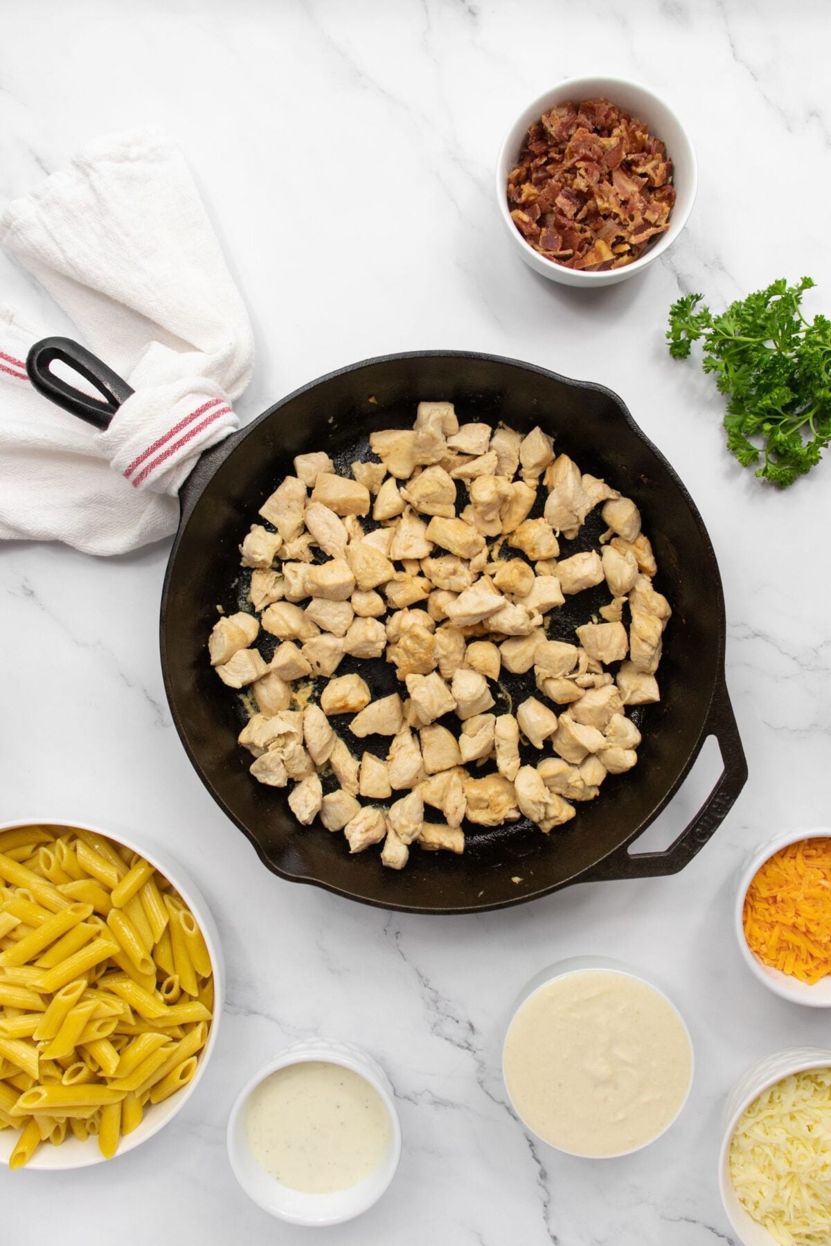 A cast iron skillet with cooked chicken pieces is surrounded by bowls of cooked penne pasta, shredded cheese, chopped bacon, white sauce, ranch dressing, and fresh parsley on a marble countertop.