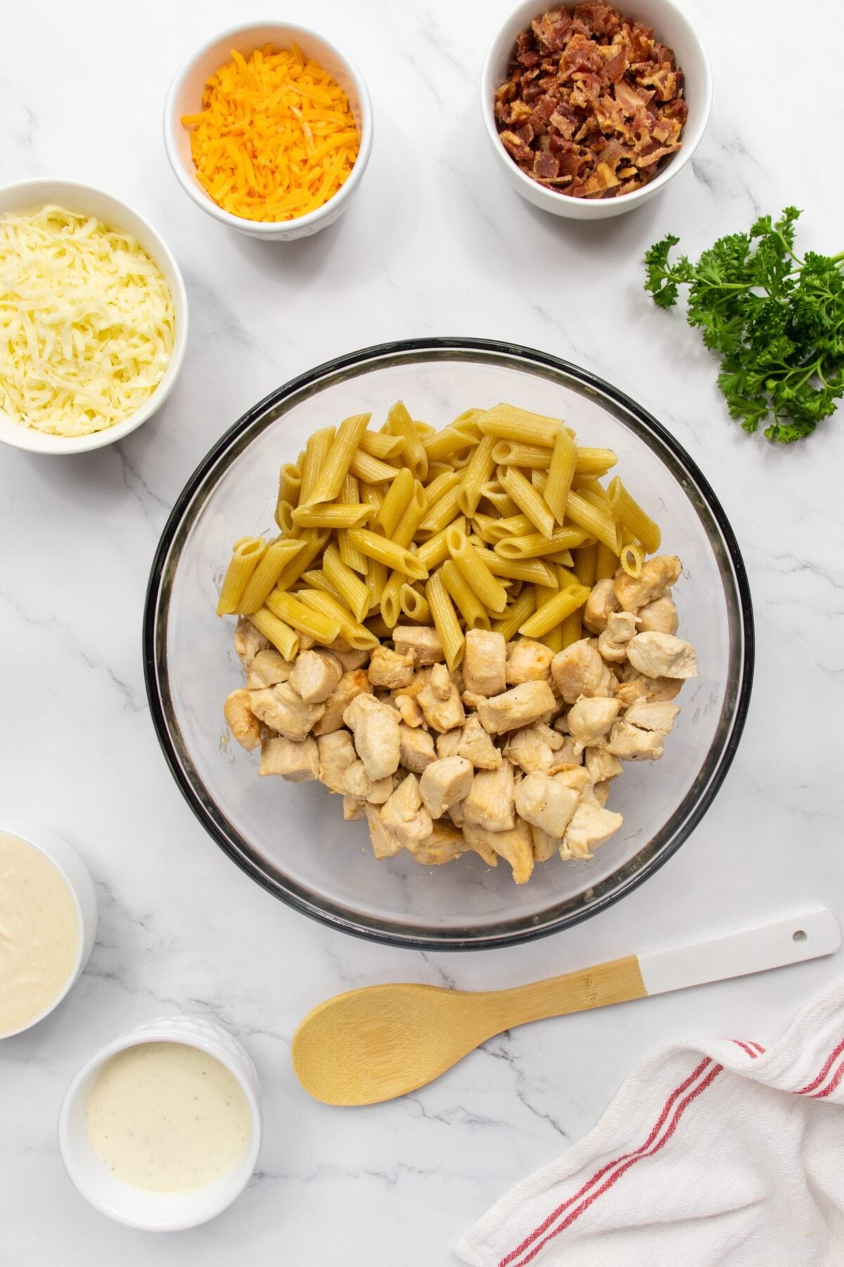 A glass bowl with cooked penne pasta and diced chicken on a marble countertop, surrounded by small bowls of shredded cheese, crumbled bacon, sauce, and fresh parsley, with a wooden spoon and a striped towel nearby.