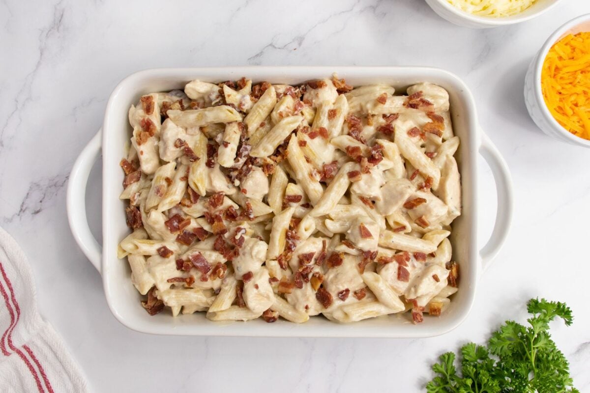A white baking dish filled with creamy penne pasta topped with chopped bacon, set on a marble surface. Small bowls of shredded cheese and fresh parsley are nearby.