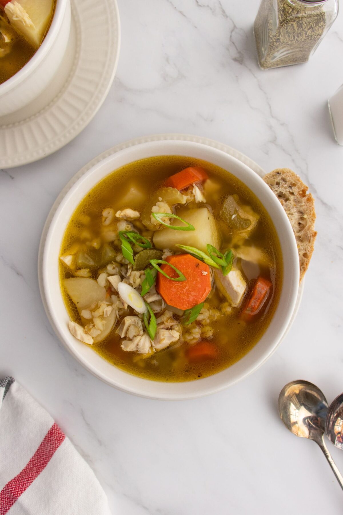 A bowl of vegetable and chicken soup with carrots, potatoes, and green onions, served with a slice of bread on a marble surface. A spoon, napkin, and pepper shaker are nearby.