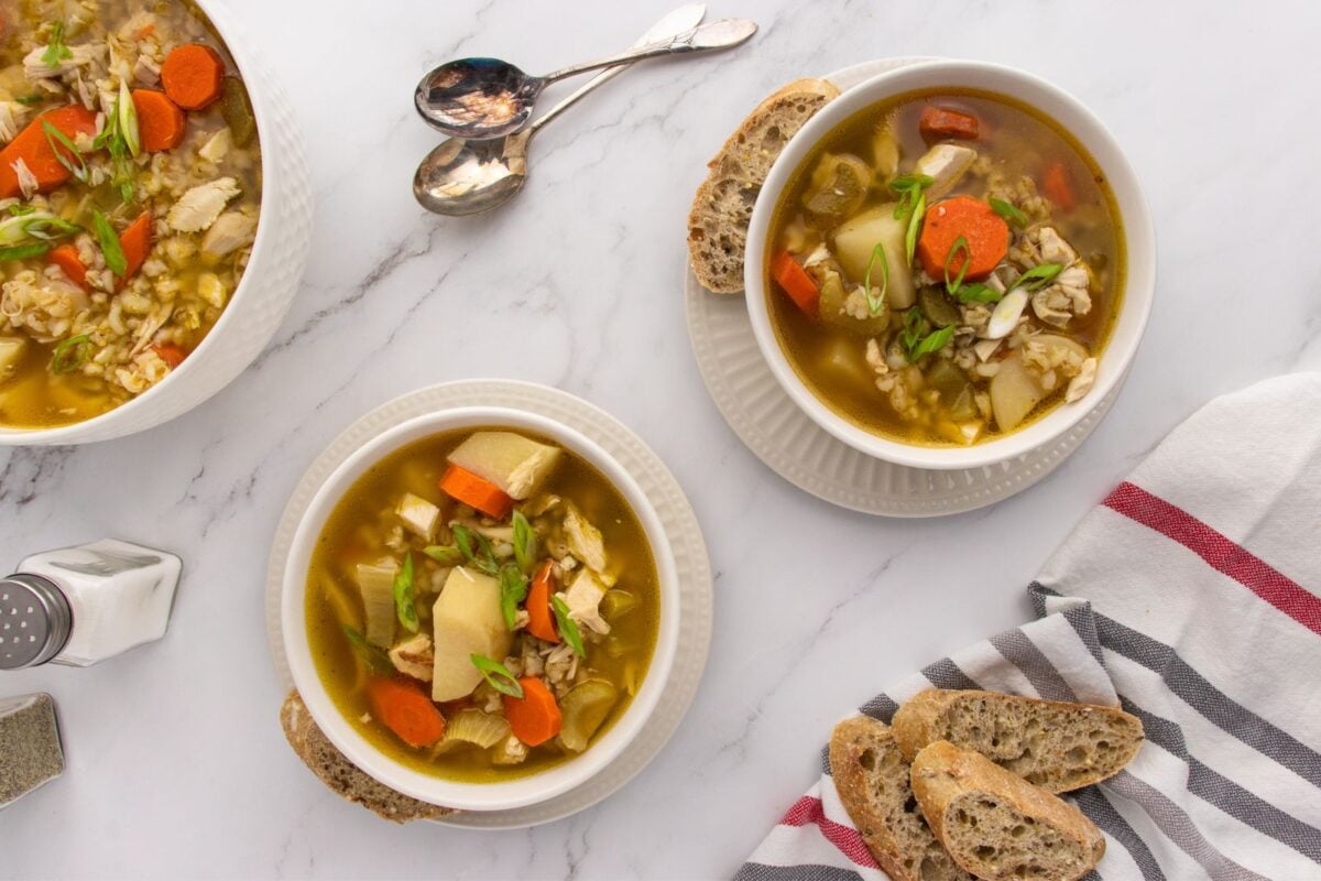 Three bowls of vegetable soup with carrots, potatoes, and chicken, garnished with bread slices. Two spoons, a salt shaker, and a striped napkin are on a white marble surface.