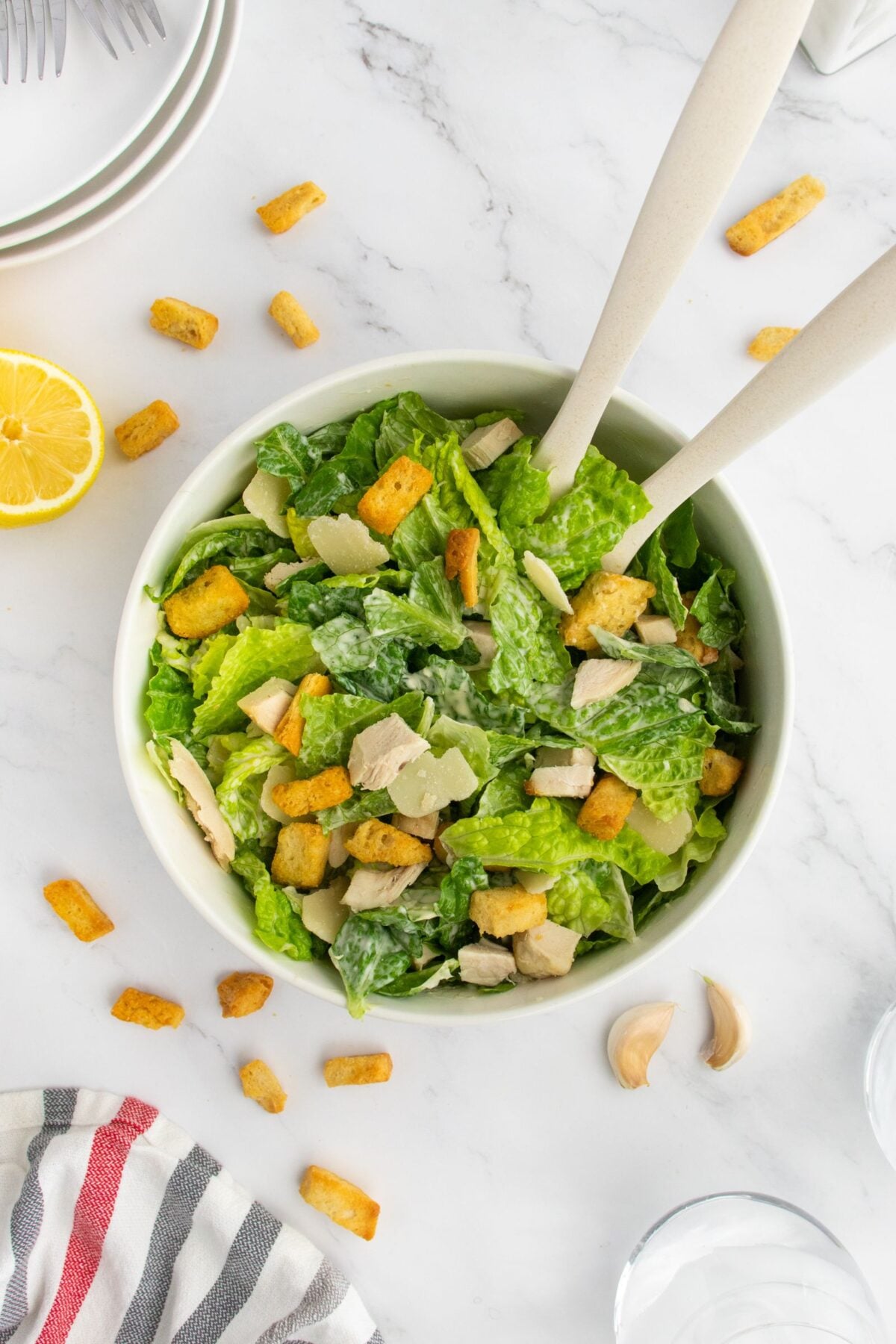 A bowl of Caesar salad with romaine lettuce, croutons, chicken, and parmesan, with two serving utensils. Scattered croutons, garlic cloves, a lemon half, and plates are nearby on a white marble surface.