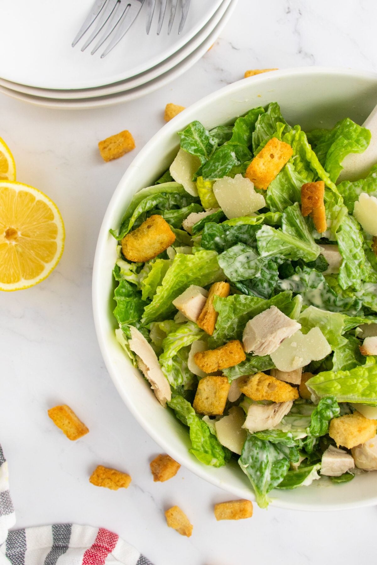 A bowl of Caesar salad with romaine lettuce, grilled chicken, croutons, and shaved parmesan on a white surface, with lemon halves, scattered croutons, plates, and forks nearby.