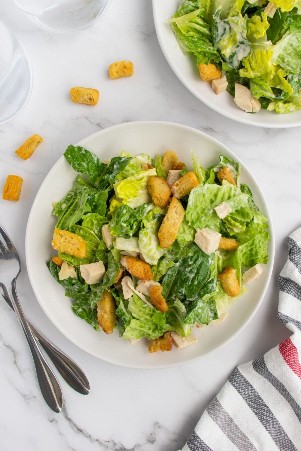 A white plate filled with Caesar salad, including romaine lettuce, croutons, and chicken pieces, sits on a marble surface near a striped napkin, two forks, and scattered croutons. Another salad plate is partly visible.