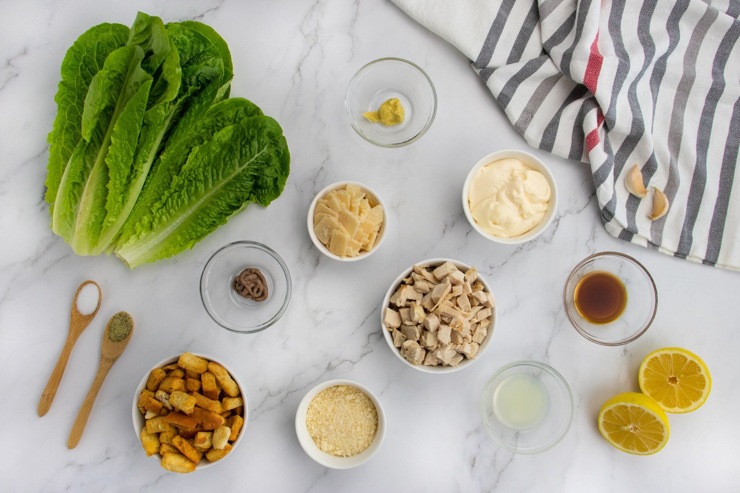 Ingredients for Caesar salad arranged on a marble surface: romaine lettuce, croutons, anchovy, mustard, parmesan, grilled chicken, Caesar dressing, lemon halves, garlic cloves, and seasonings with a striped towel.