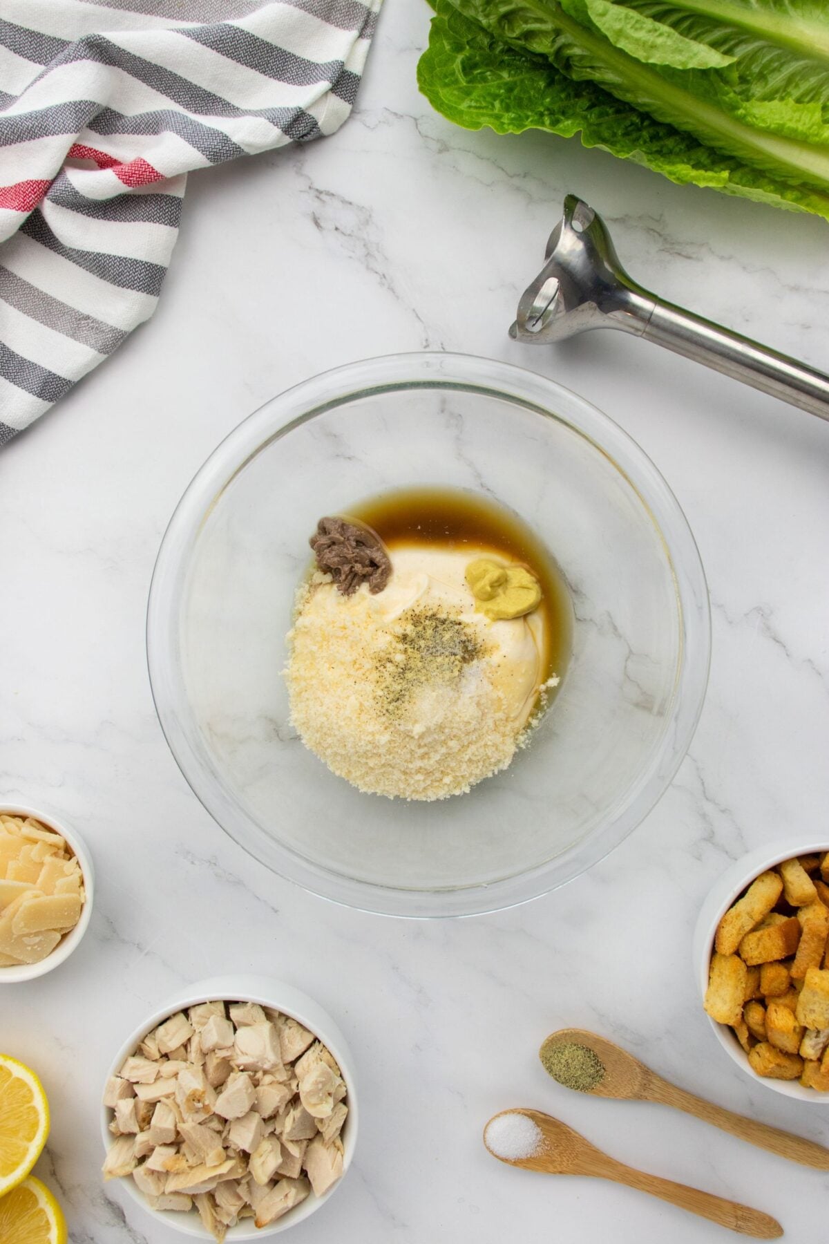A glass bowl with Caesar salad dressing ingredients, including mayo, parmesan, mustard, and Worcestershire sauce, sits on a marble countertop surrounded by lettuce, chicken, croutons, lemon, and kitchen tools.