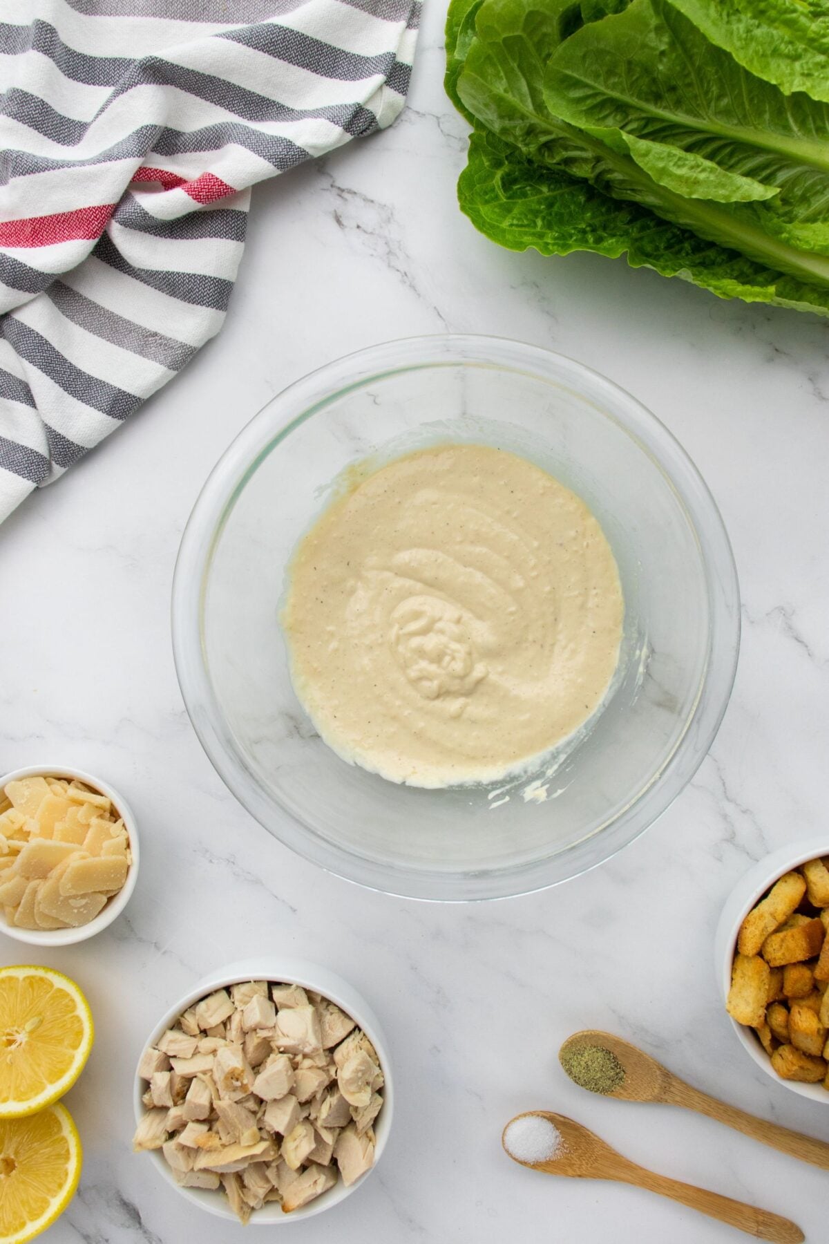 A glass bowl with creamy dressing is on a marble surface, surrounded by romaine lettuce, croutons, lemon halves, sliced parmesan, diced chicken, pepper, and salt. A striped towel is in the top left corner.