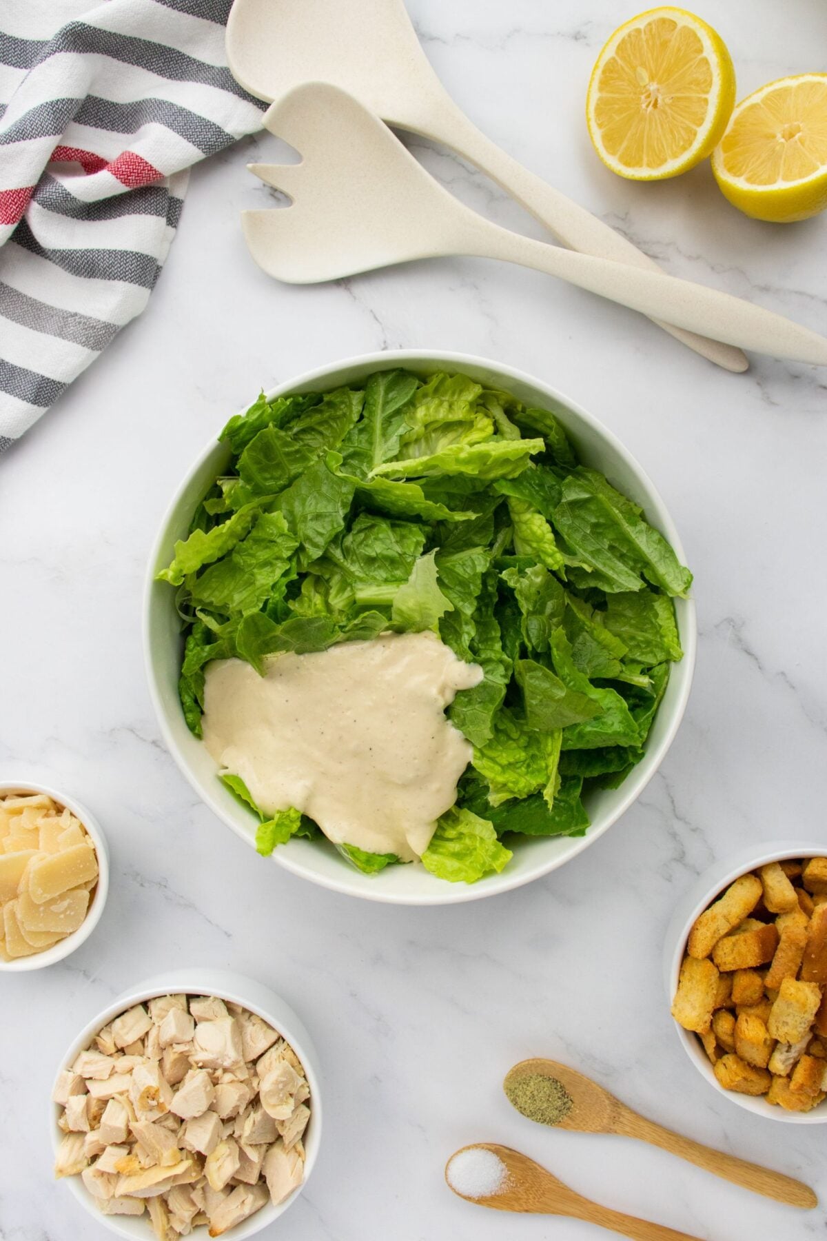 A bowl of romaine lettuce with Caesar dressing sits on a marble surface, surrounded by small bowls of croutons, chicken, parmesan, a striped towel, wooden salad utensils, lemon halves, and wooden spoons with spices.