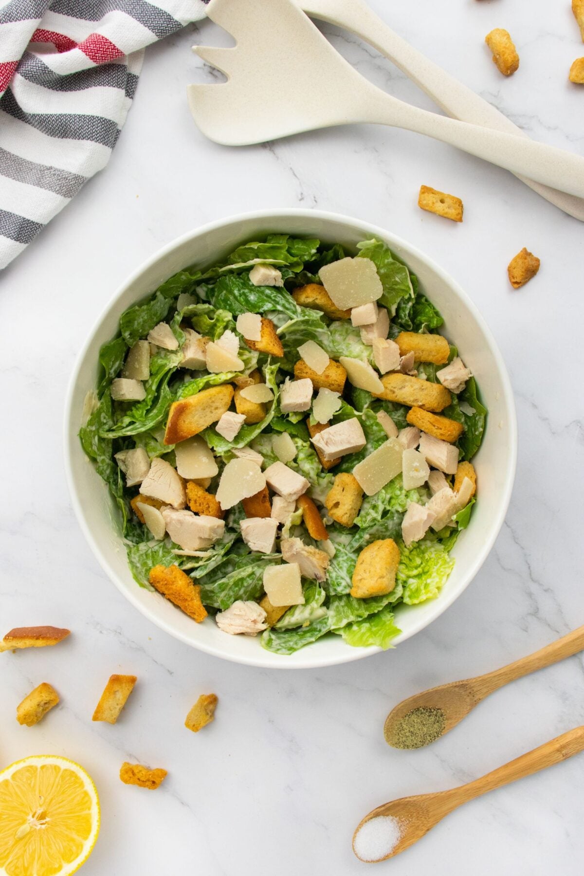 A bowl of Caesar salad with chopped chicken, croutons, and Parmesan cheese on a marble surface, surrounded by wooden utensils, a striped towel, lemon, and spice-filled wooden spoons.