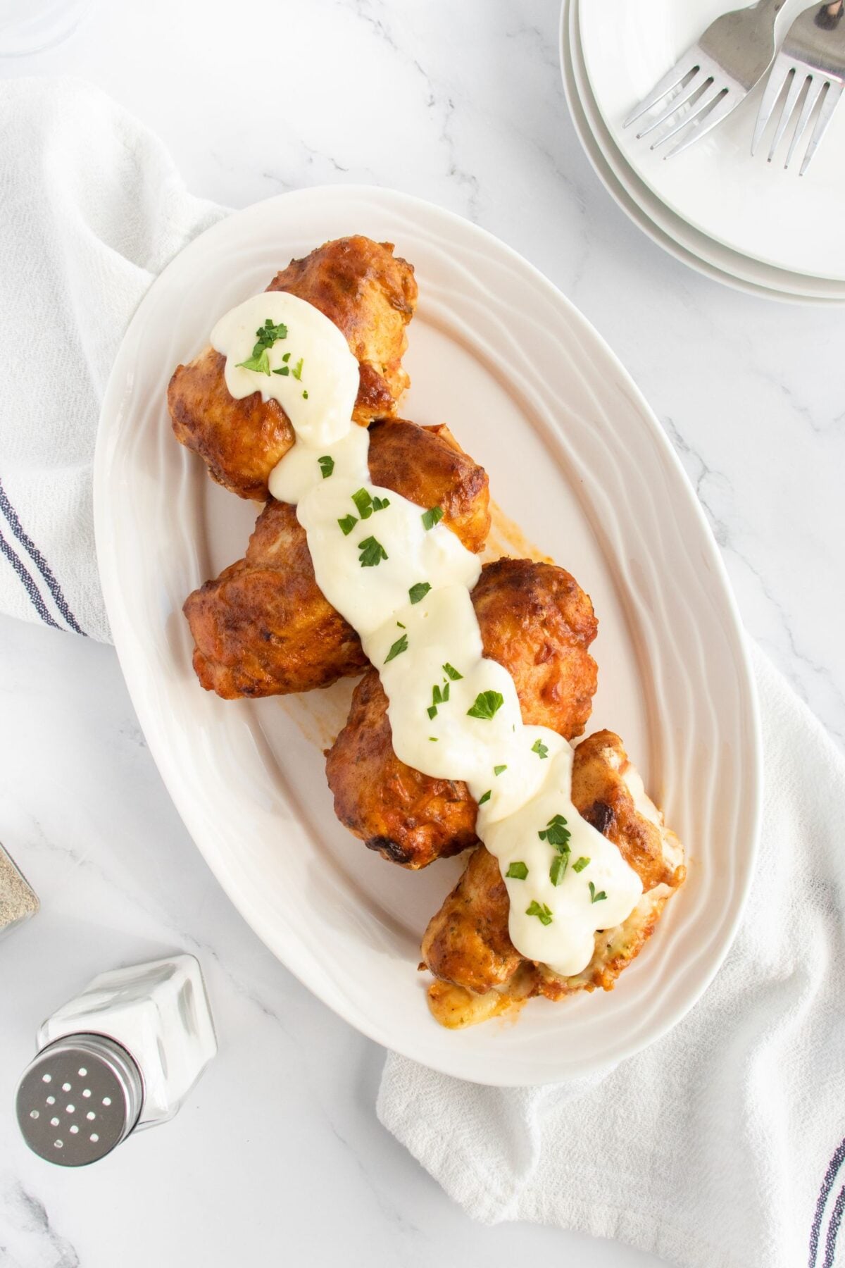 Four pieces of fried chicken topped with white creamy sauce and chopped herbs, arranged on a white oval plate, with a salt shaker and stacked plates with forks nearby.