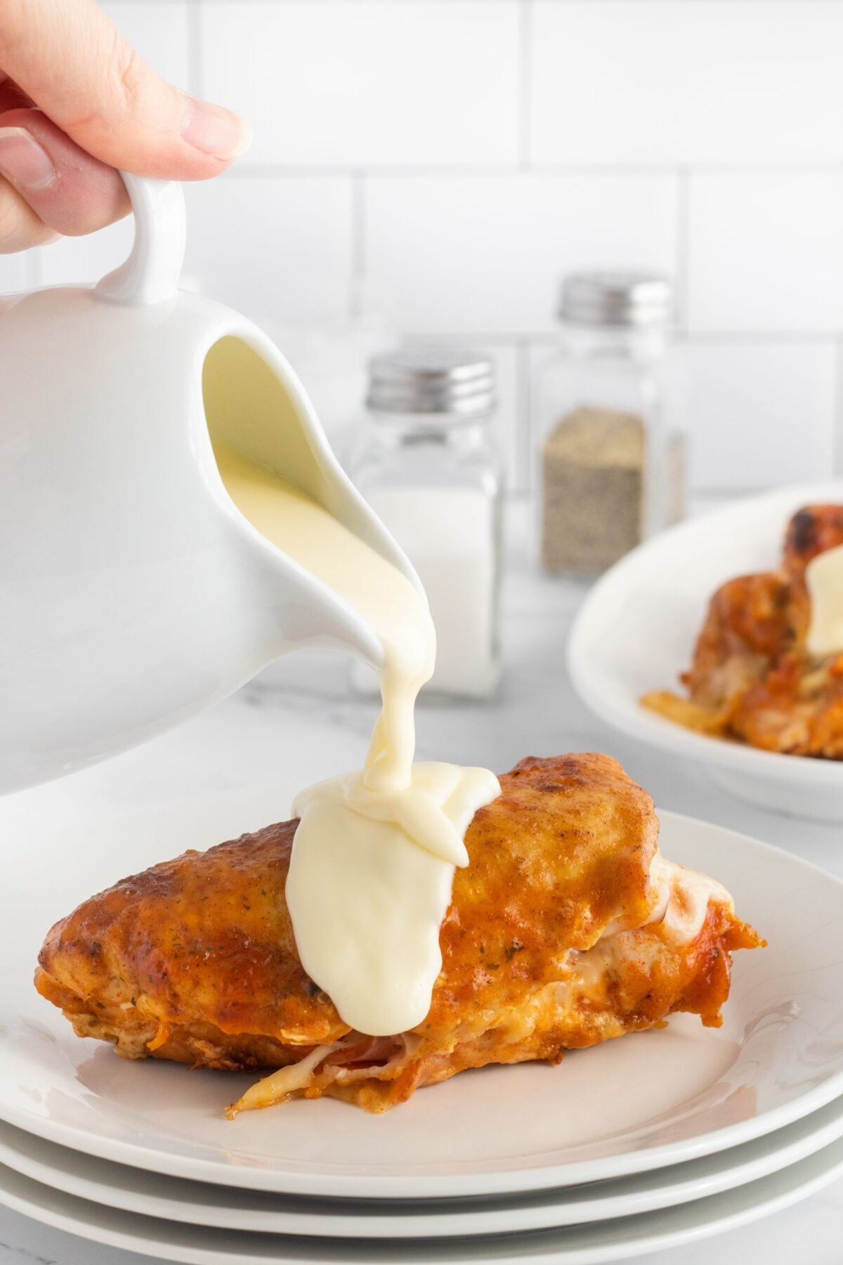 A hand pours creamy white sauce from a small pitcher onto a stuffed, baked chicken breast on a white plate. In the background, more chicken and salt and pepper shakers are visible.