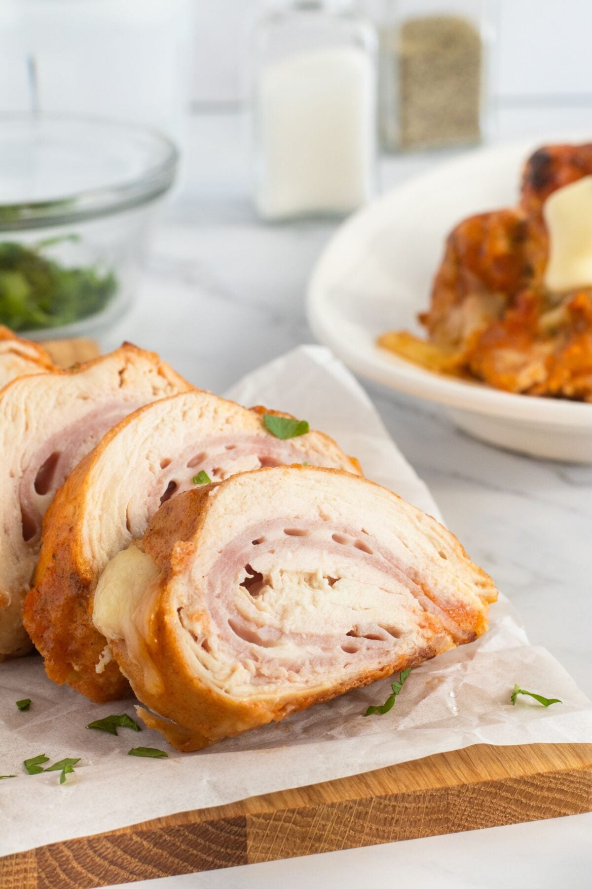 Three slices of stuffed chicken breast, rolled with layers of ham and cheese, are served on parchment paper atop a wooden board. A bowl of greens and a white plate with more stuffed chicken are in the background.