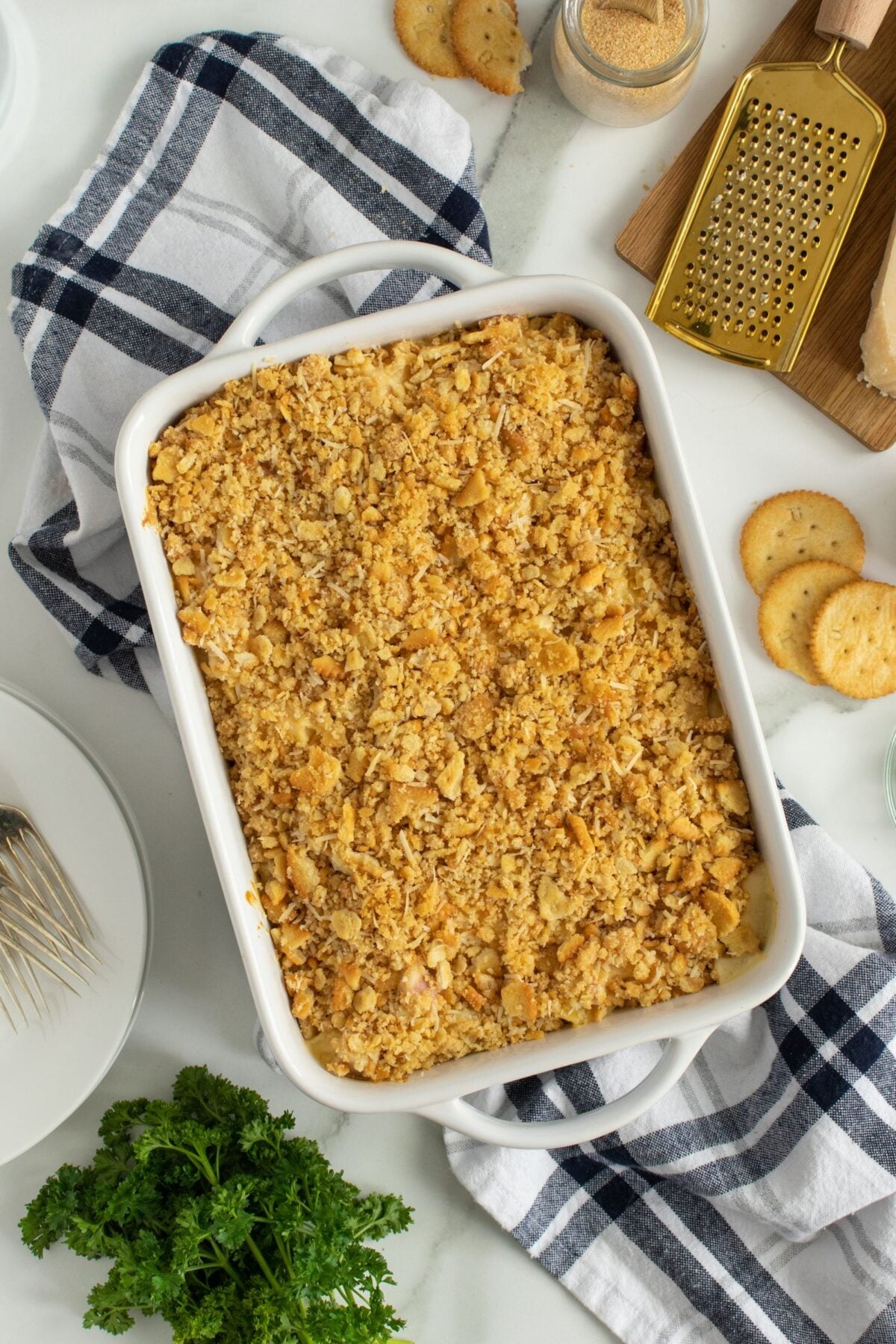 A white baking dish filled with a casserole topped with a golden, crumbly cracker crust sits on a striped kitchen towel, surrounded by parsley, whole crackers, plates, and kitchen utensils.