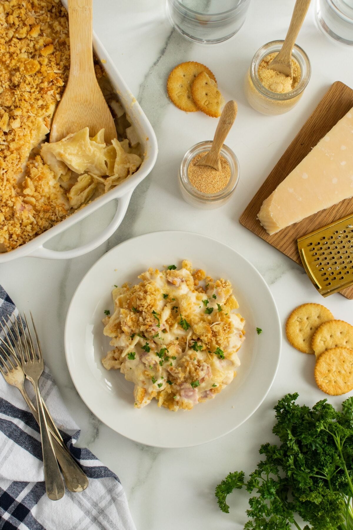 A serving of cheesy casserole topped with crushed crackers is plated next to a baking dish with more casserole. Surrounding are crackers, grated cheese, parsley, utensils, and a checked napkin on a white marble surface.