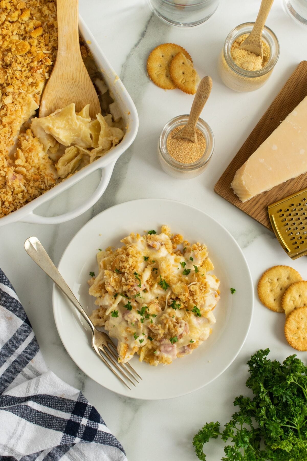 A plate of creamy baked casserole topped with crushed crackers sits beside a baking dish of the same casserole. Ingredients like crackers, grated cheese, and parsley are nearby on a marble countertop.